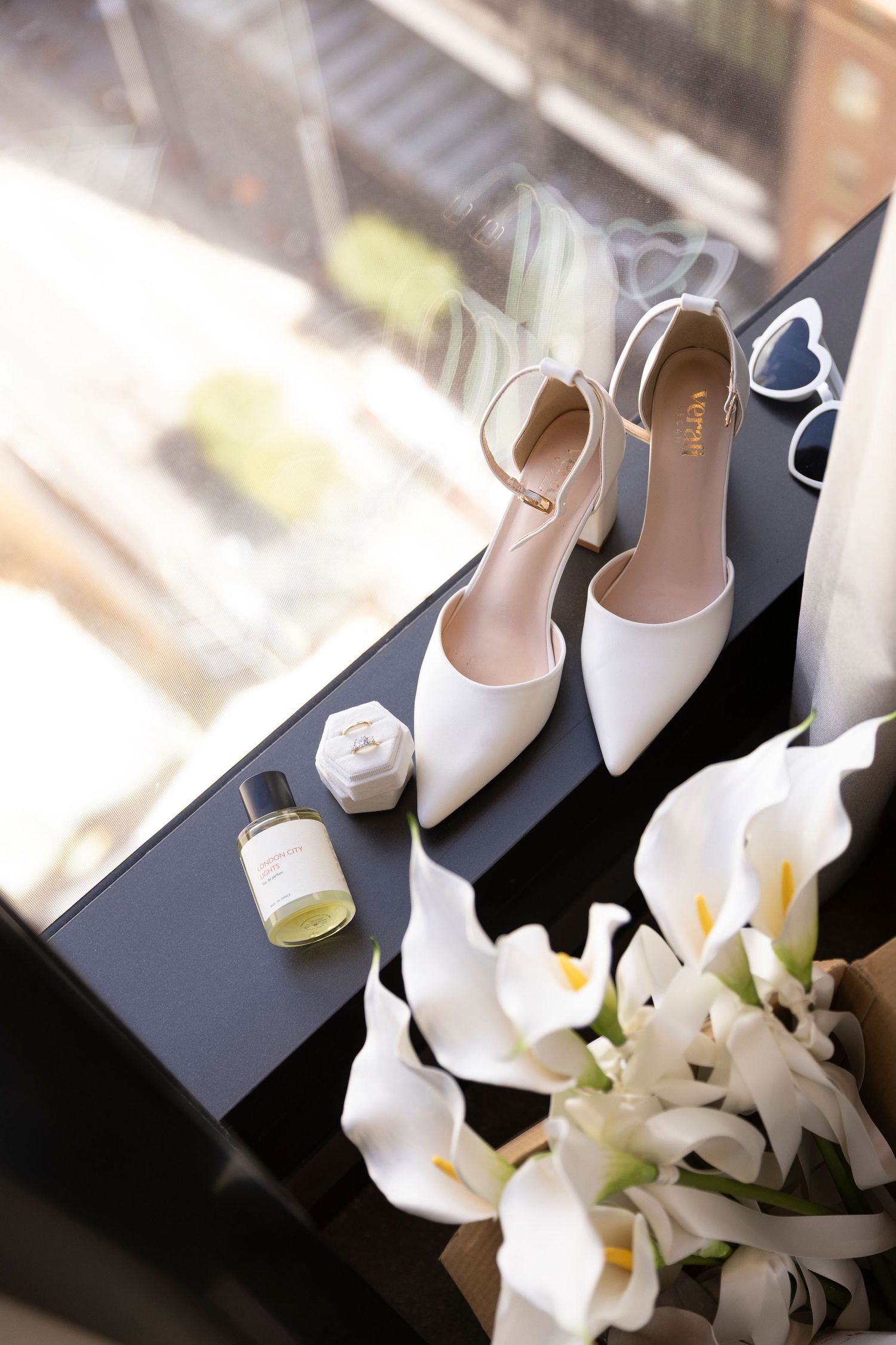 White bridal heels, perfume, heart sunglasses, and calla lily bouquet arranged on a dark surface near a window.