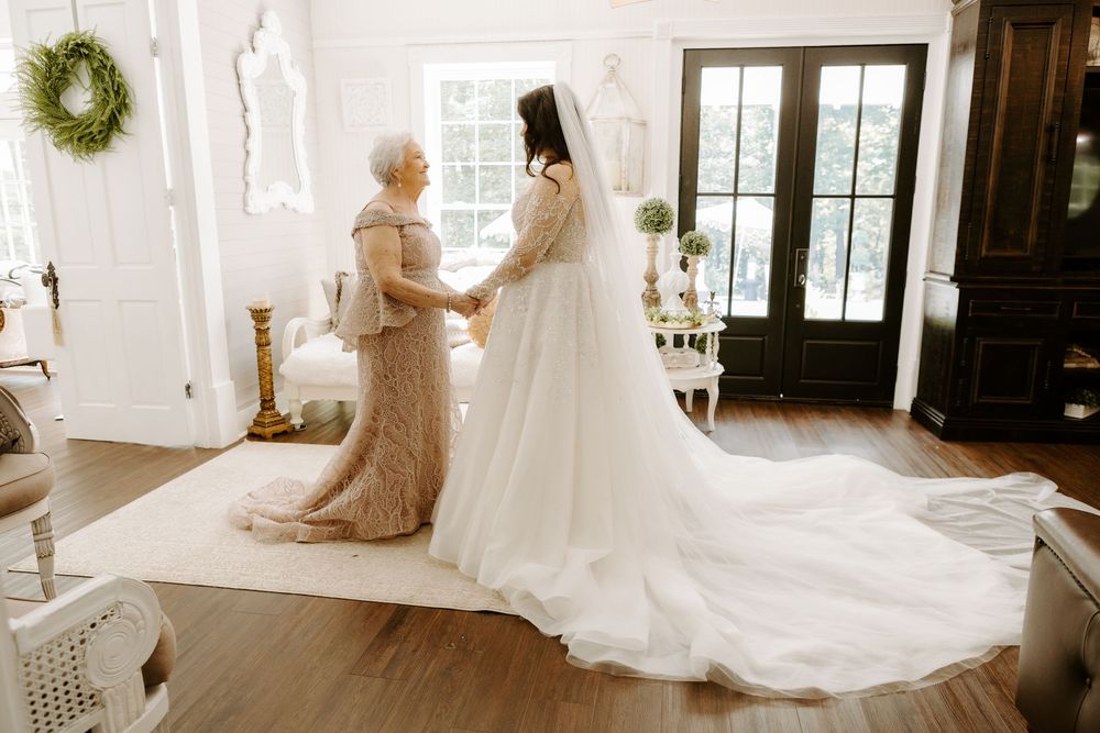 A bride in a white gown with long train holds hands with an older woman in a gold dress inside an elegant room.