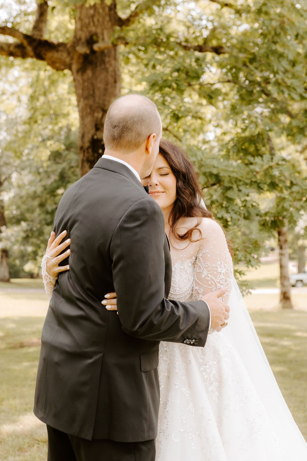 Bride and groom share a tender embrace outdoors under a large tree on their wedding day.