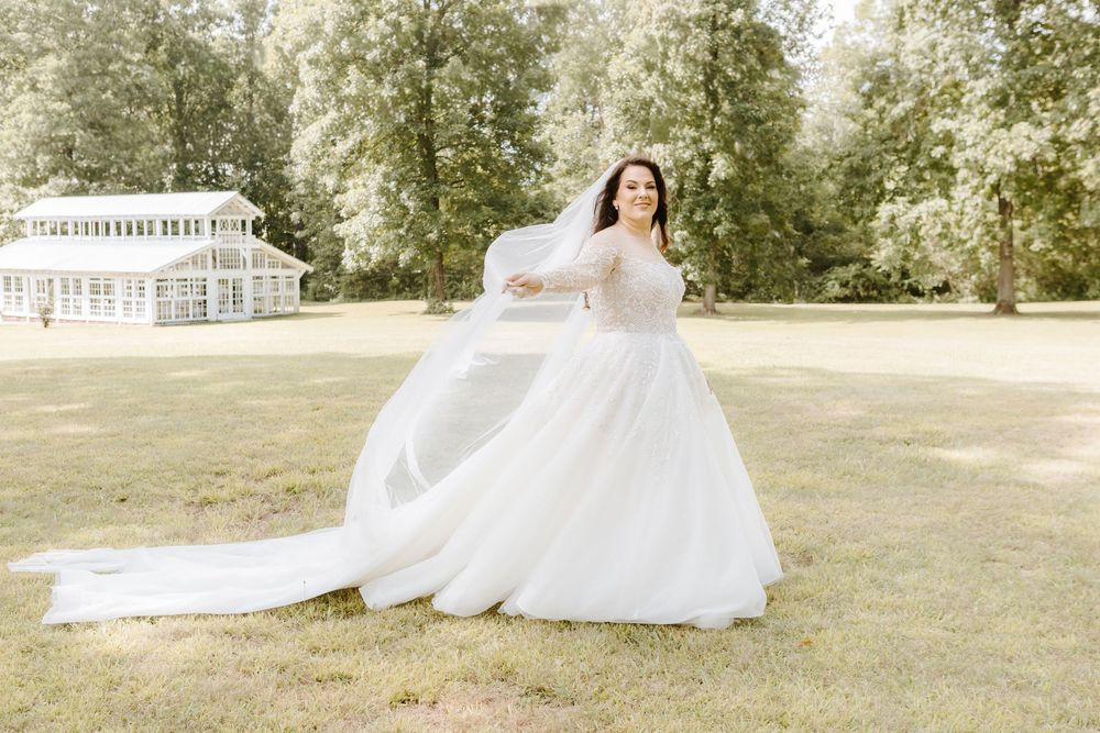 Bride in a flowing white ball gown twirling outdoors in a lush green park with a glass greenhouse backdrop.