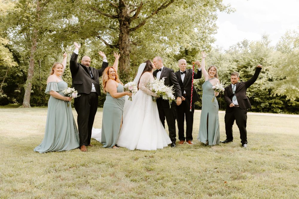 Wedding party celebrates outdoors in sage green bridesmaid dresses and black tuxedos, cheering with bride and groom on green lawn.