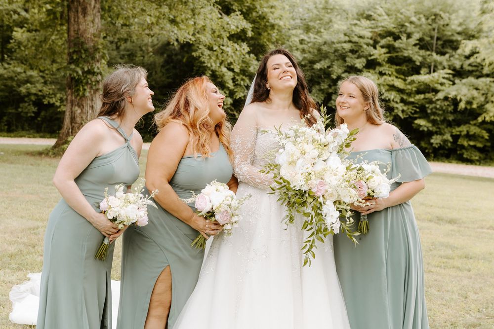 Bride in white gown laughing with three bridesmaids in sage green dresses holding white bouquets outdoors.