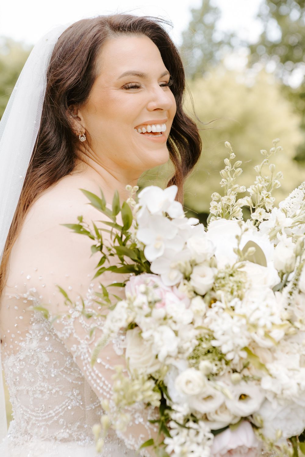 Smiling bride in beaded lace gown holding lush white floral bouquet with greenery, wearing veil and pearl earrings.
