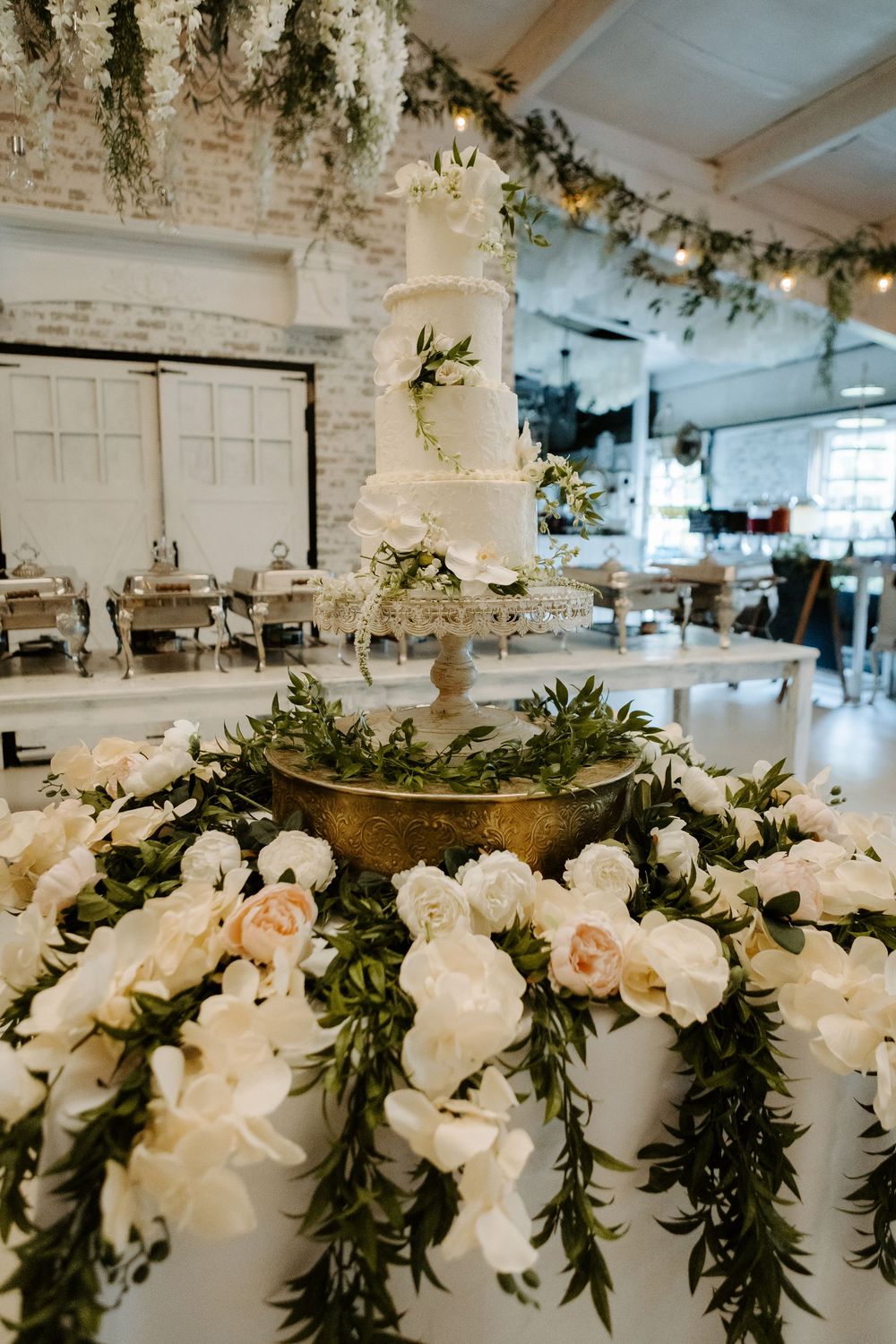 Elegant white tiered wedding cake displayed on a gold stand surrounded by lush white floral arrangements and greenery garlands.