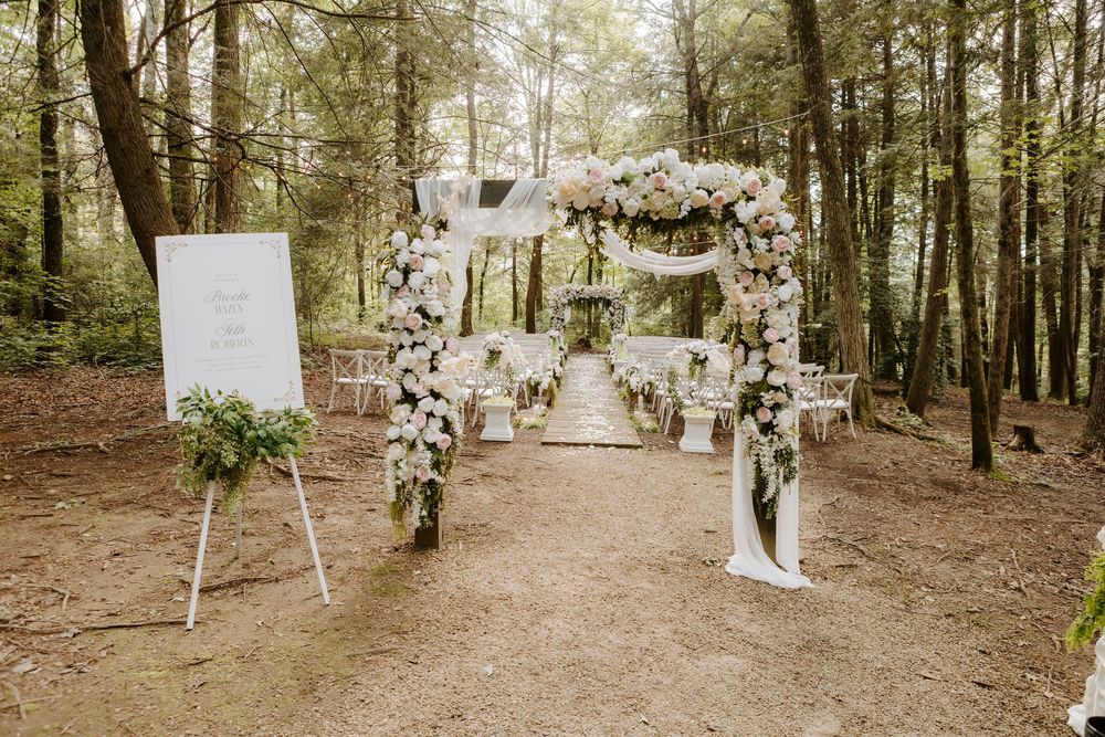 Elegant outdoor forest wedding ceremony with floral arch, white chairs, and welcome sign amid tall trees.