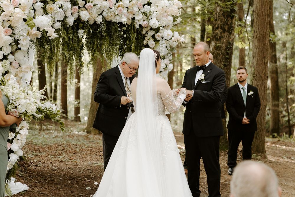 Outdoor woodland wedding ceremony with bride in white gown, groom in black tuxedo, and officiant under floral arch.