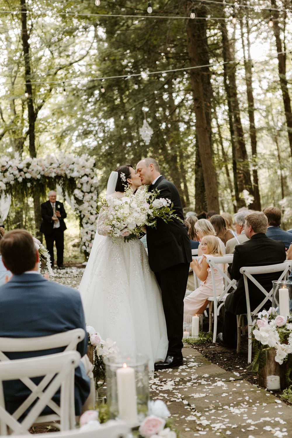Bride and groom share first kiss at outdoor forest wedding ceremony with white floral arch and rose petal aisle.