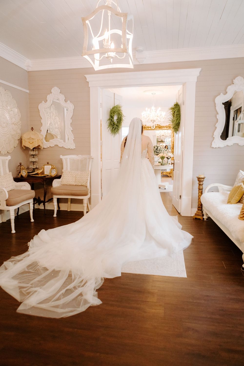 Bride in a stunning long white gown with flowing veil stands in an elegant bridal suite doorway.