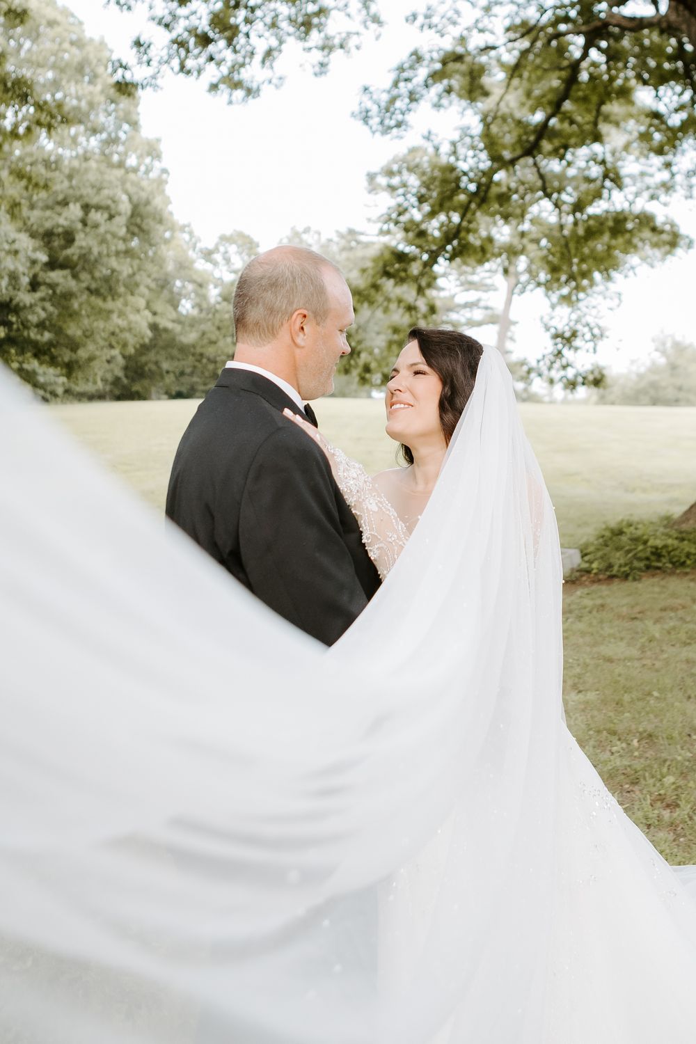 Bride and groom share a romantic moment outdoors with veil flowing in the breeze.
