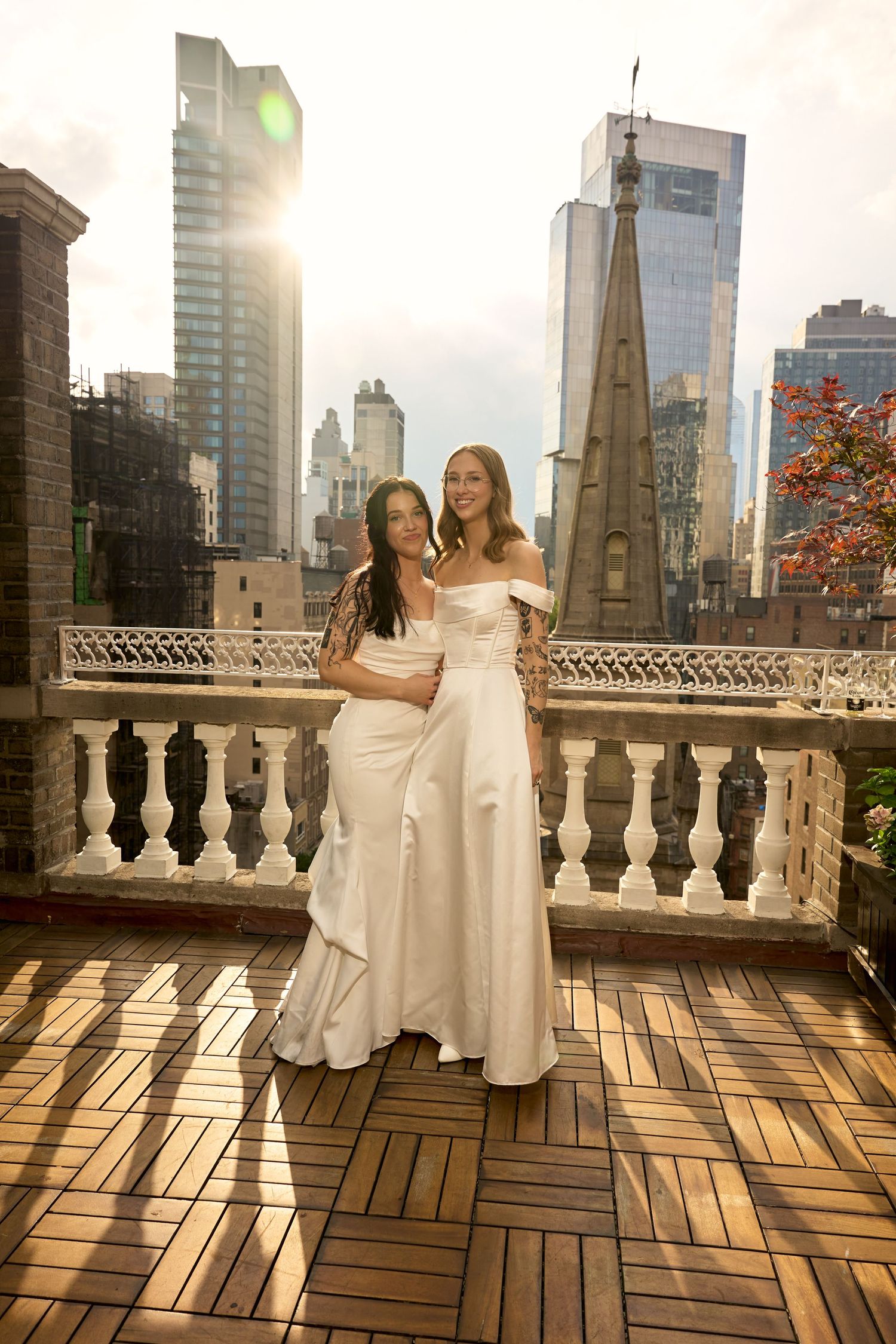 Two brides in white gowns pose on a rooftop terrace with NYC skyline backdrop at golden hour.