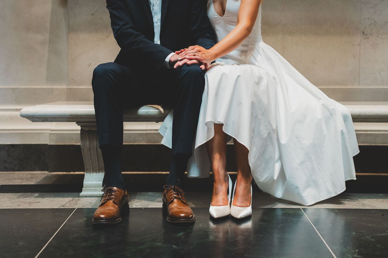 Bride and groom sitting together, wearing white heels and brown dress shoes, with wedding attire visible from waist down.