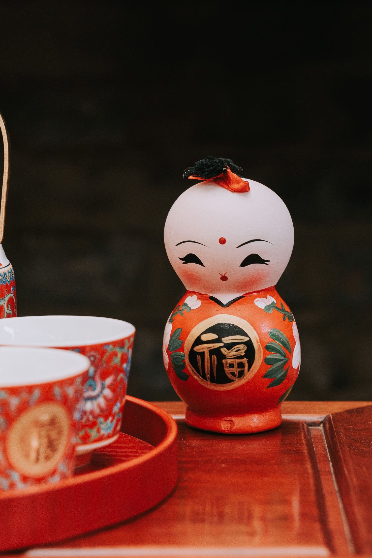 A traditional Japanese Kokeshi doll with red and green floral design sits beside red ceramic tea cups on a wooden tray.