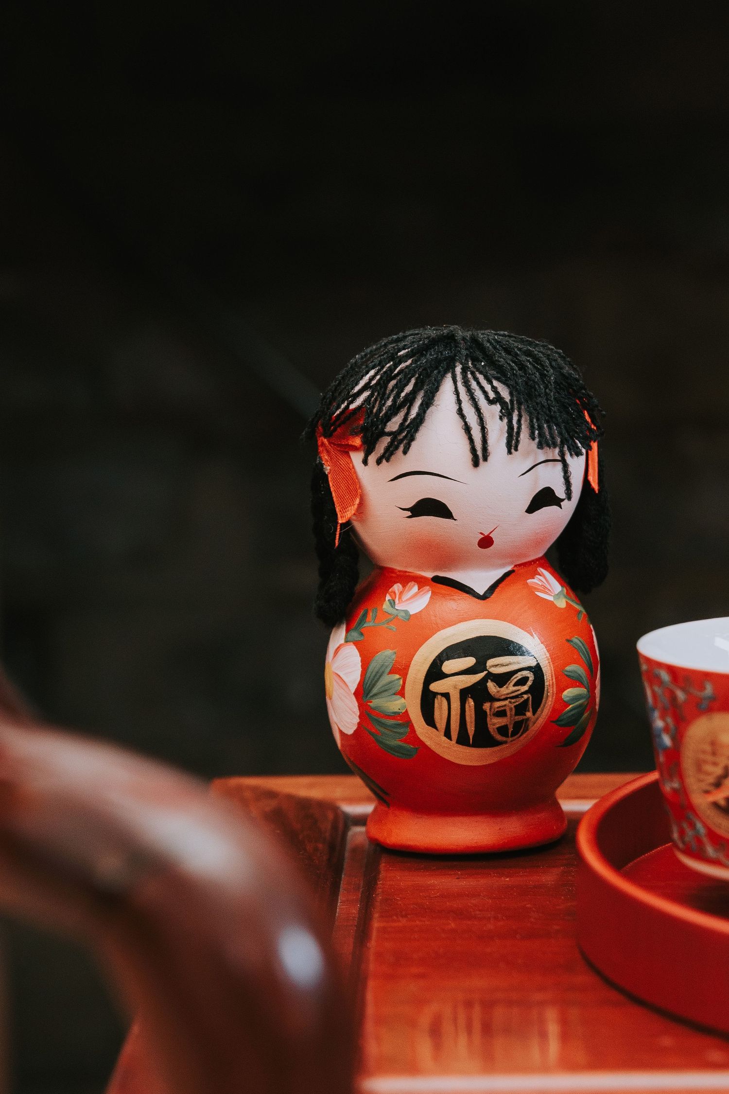 Red Japanese Kokeshi doll with black hair and kanji symbol, displayed on wooden tray with tea set.