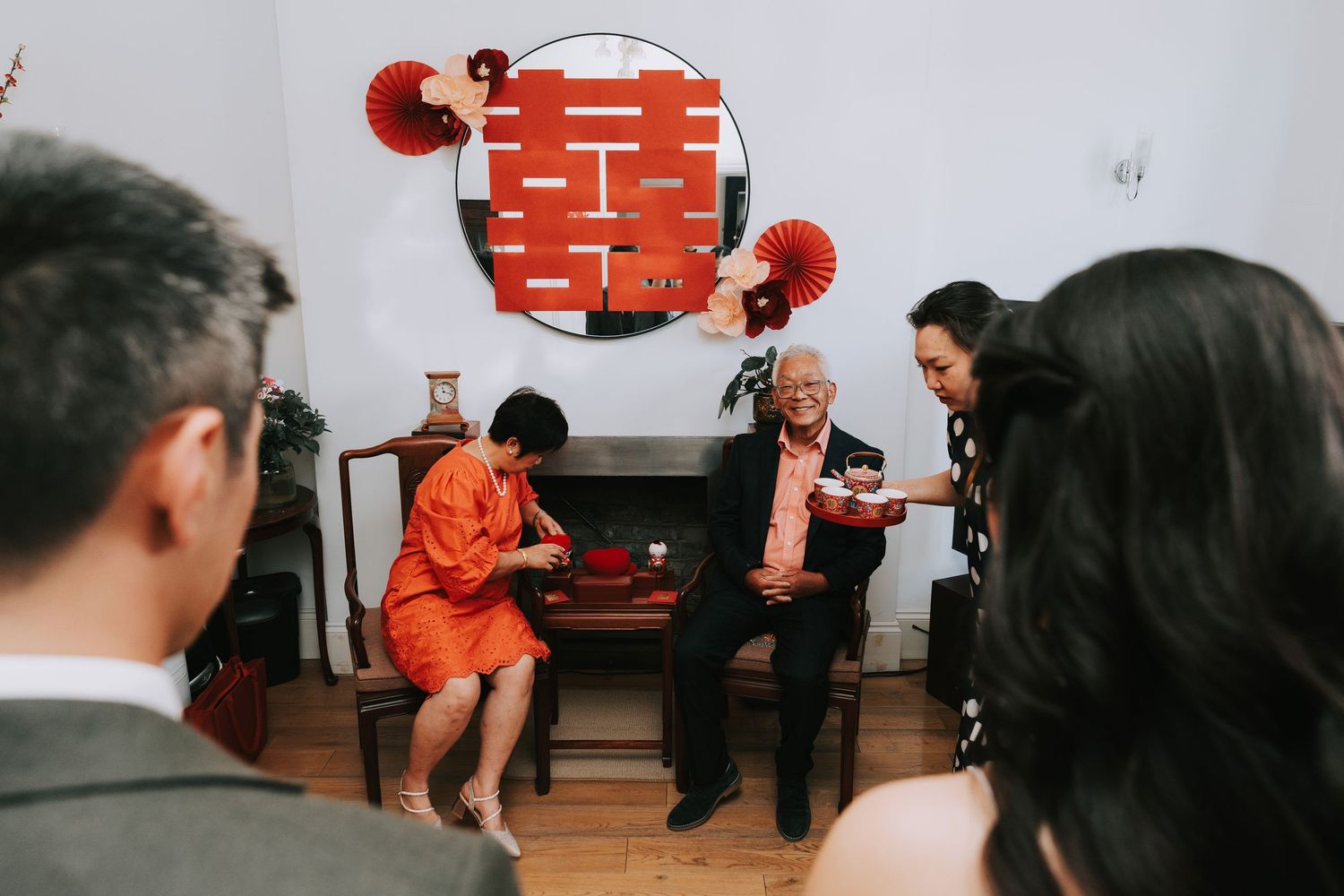Traditional Chinese wedding tea ceremony with elders seated on wooden chairs, red double happiness decorations on white wall.