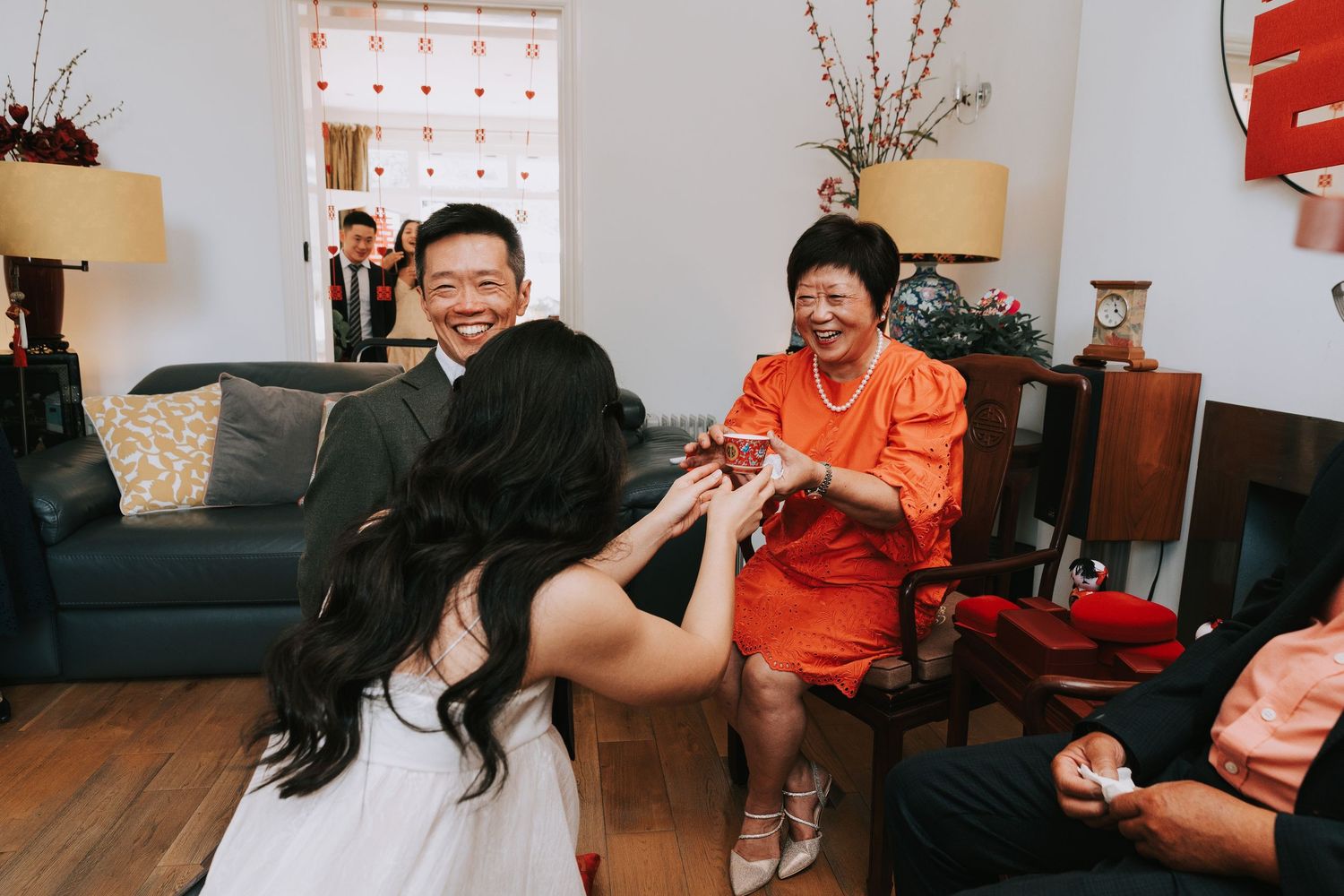 A bride offers tea to a smiling older woman in orange during a traditional Chinese tea ceremony indoors.