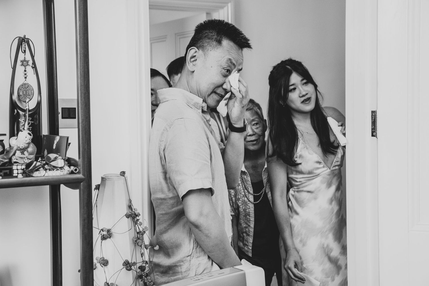 Emotional black and white photo of a man wiping tears at a wedding ceremony with a woman in a satin dress nearby.