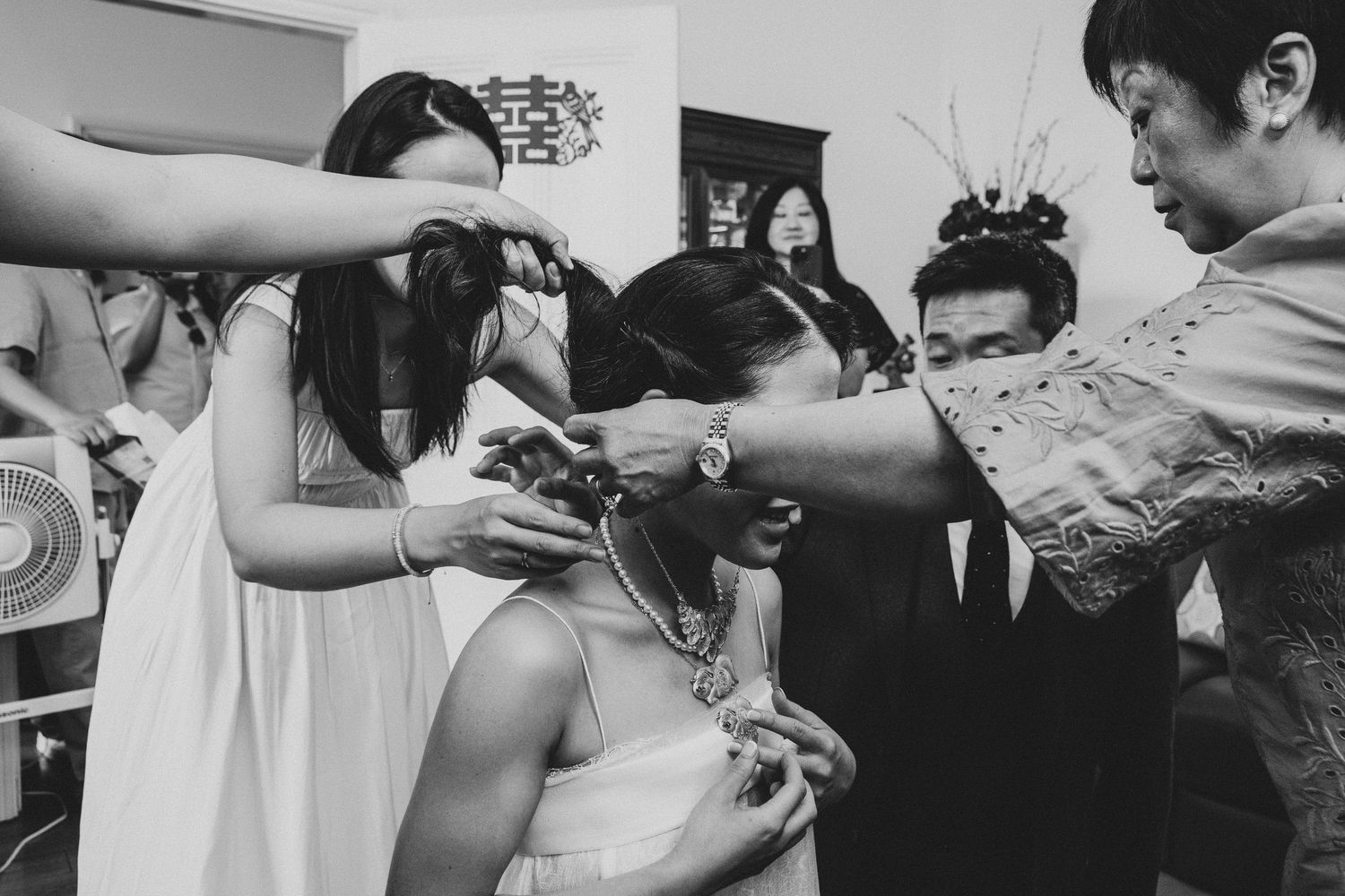 Black and white photo of a bride being helped with jewelry and hair by wedding party members indoors.