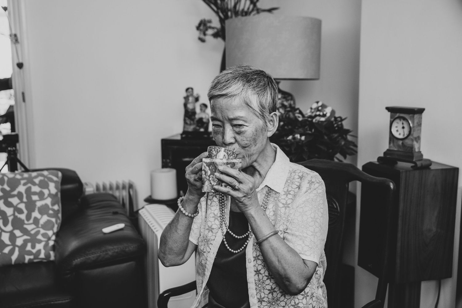 Elderly Asian woman sipping tea in a cozy living room, captured in black and white photography.