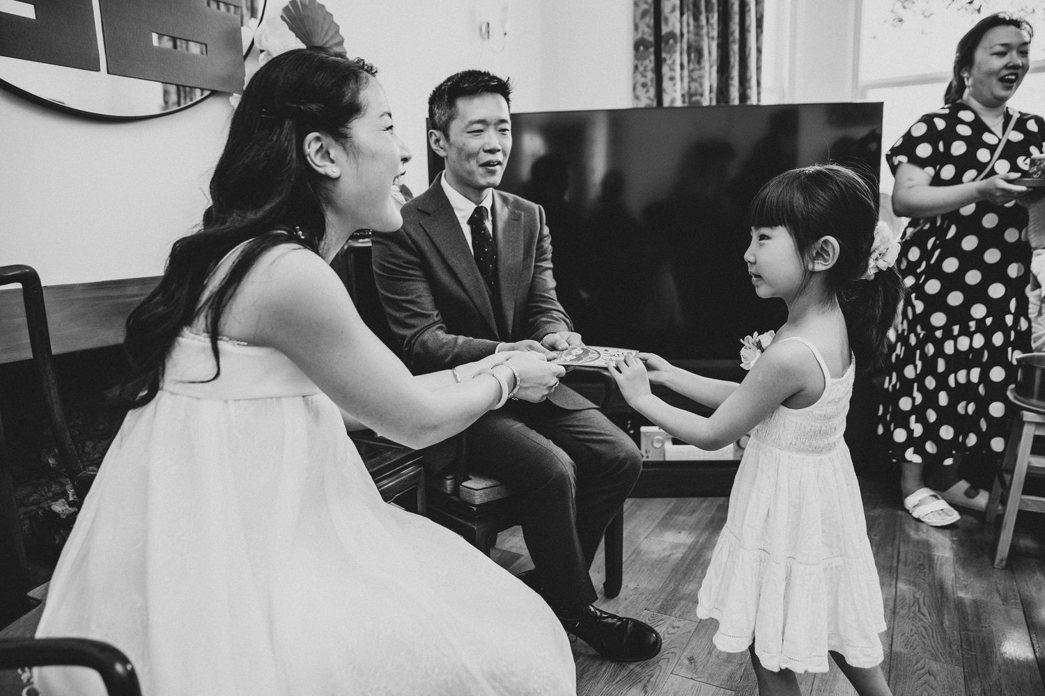 Black and white photo of a bride and groom interacting with a young flower girl at a wedding reception.