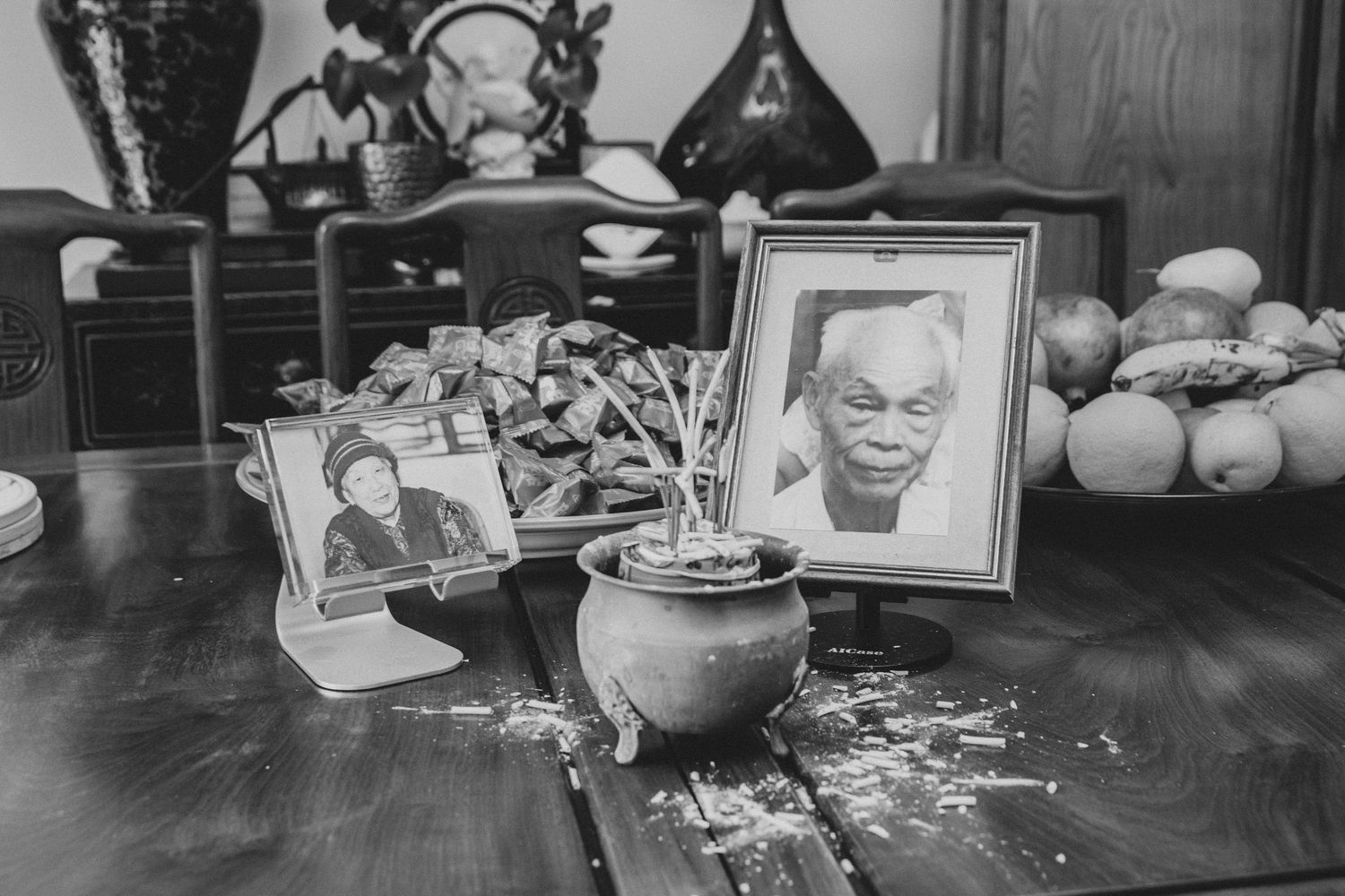 Black and white photo of a memorial altar with framed portraits of elderly people, a small plant, and fruit offerings on a wooden table.
