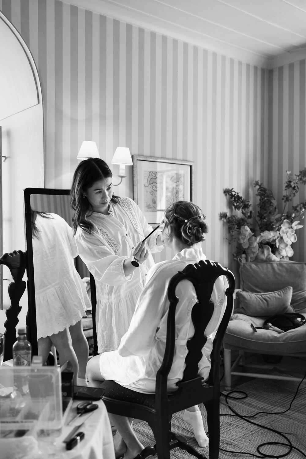 Black and white photo of a makeup artist applying makeup to a bride seated in an elegant chair before her wedding.