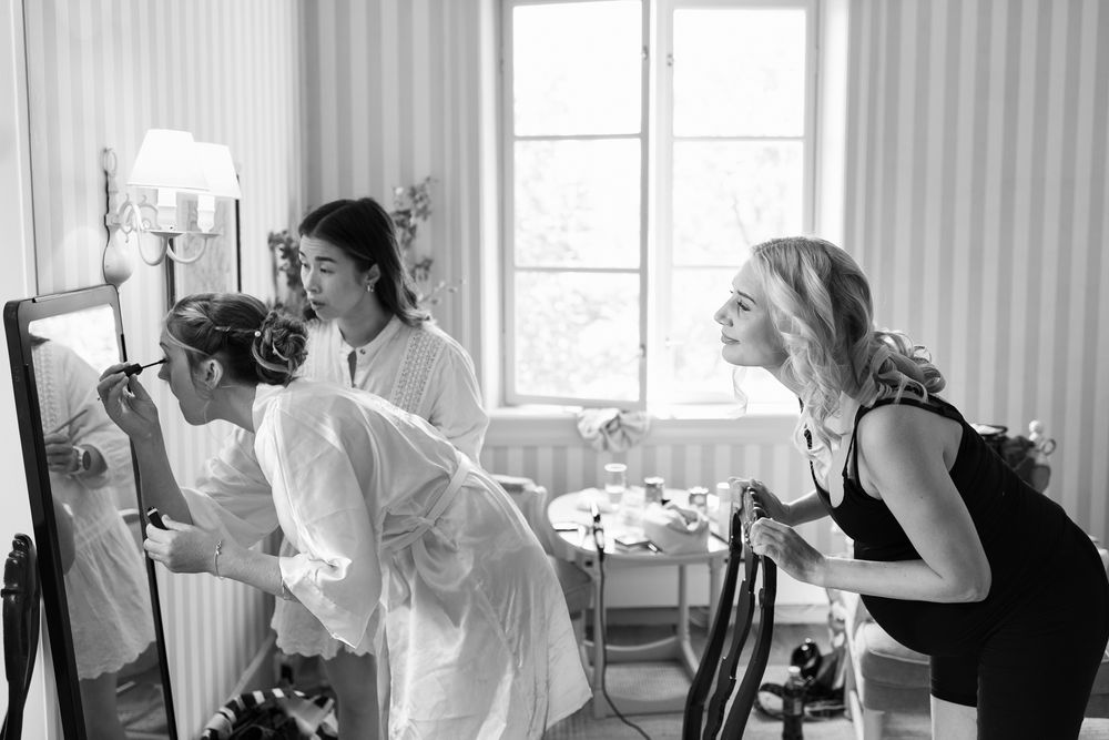 Black and white photo of bridesmaids helping bride with makeup and hair in elegant room before wedding ceremony.