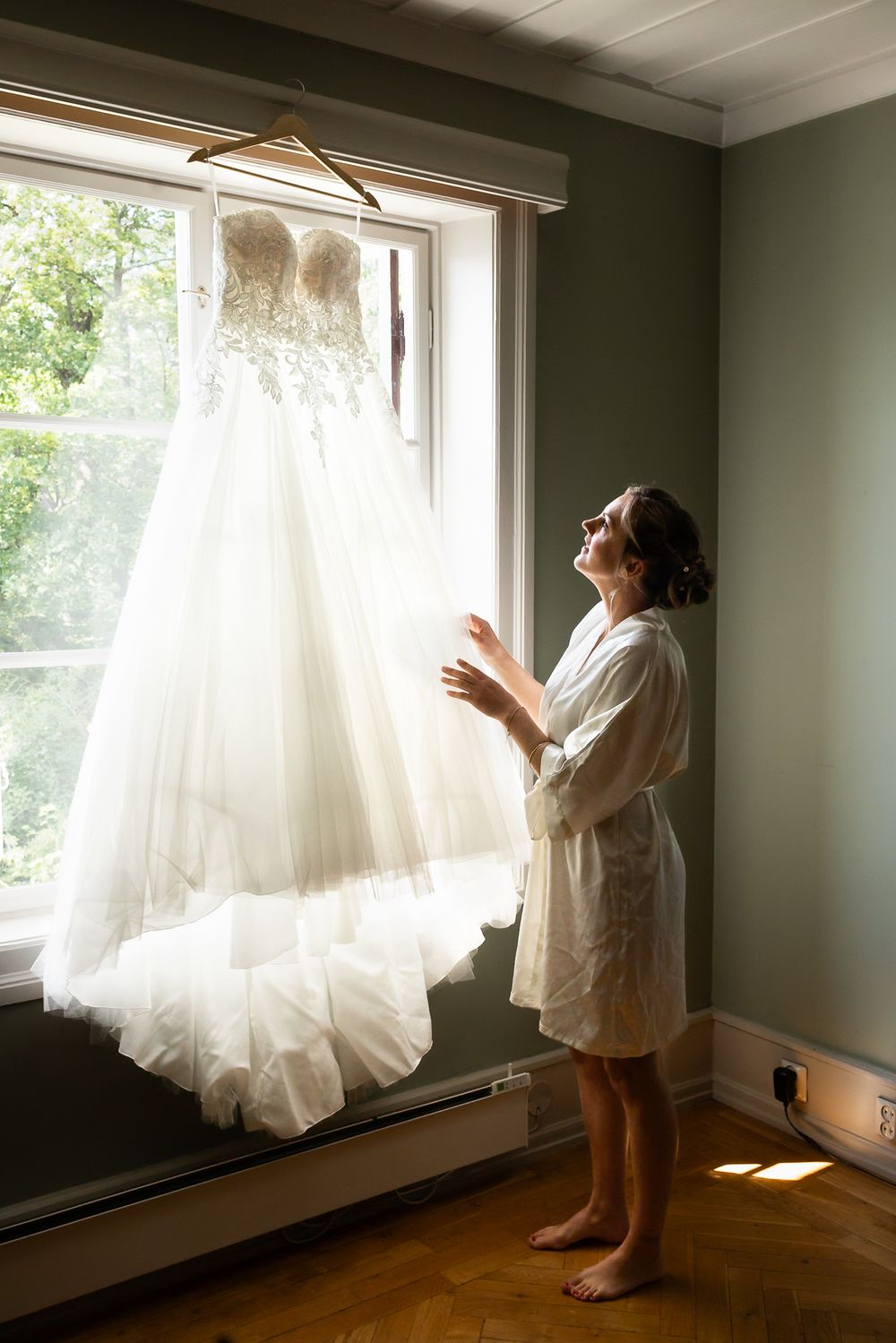 Bride in robe admiring her lace wedding gown hanging by a sunlit window on her wedding day.