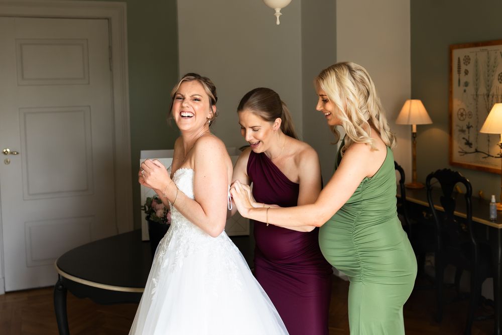 Bride laughing with two bridesmaids in purple and green dresses helping zip up her white wedding gown indoors.
