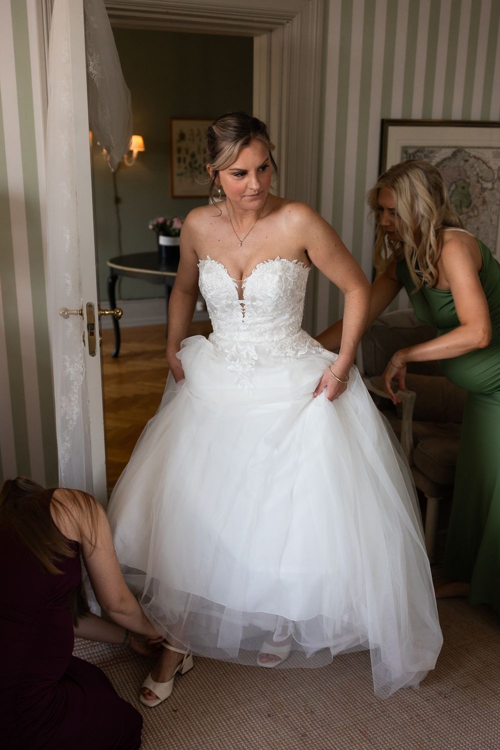 Bride in white strapless ballgown being helped by bridesmaids in elegant room before wedding ceremony.