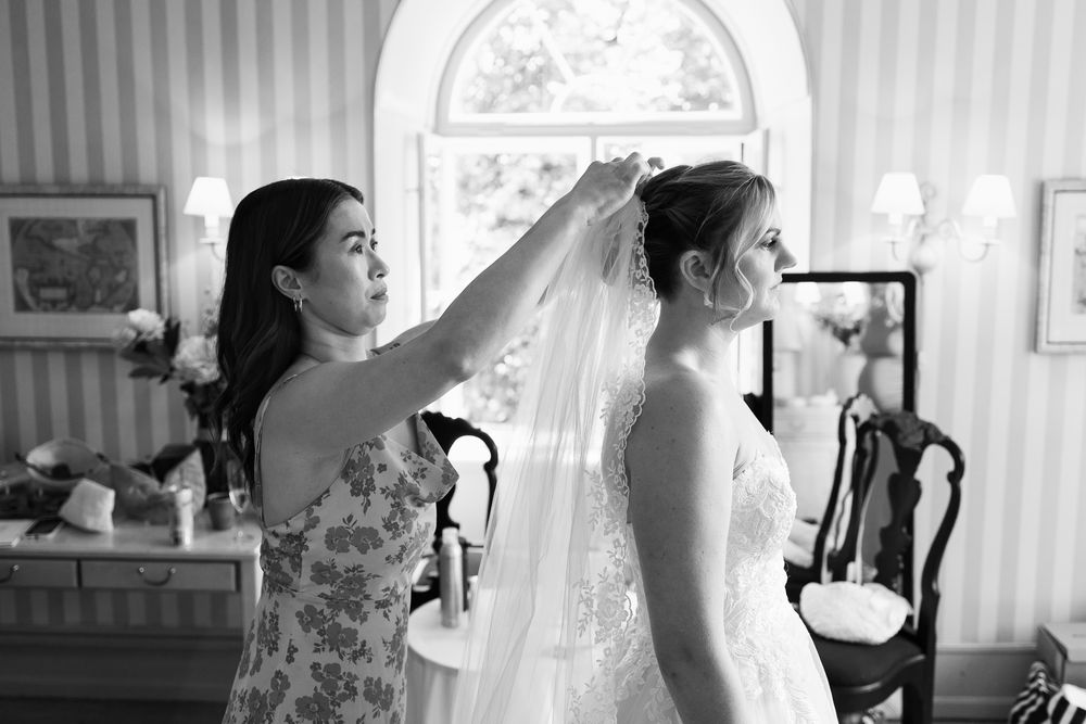 Black and white photo of a woman helping a bride adjust her veil in an elegant room before the wedding ceremony.