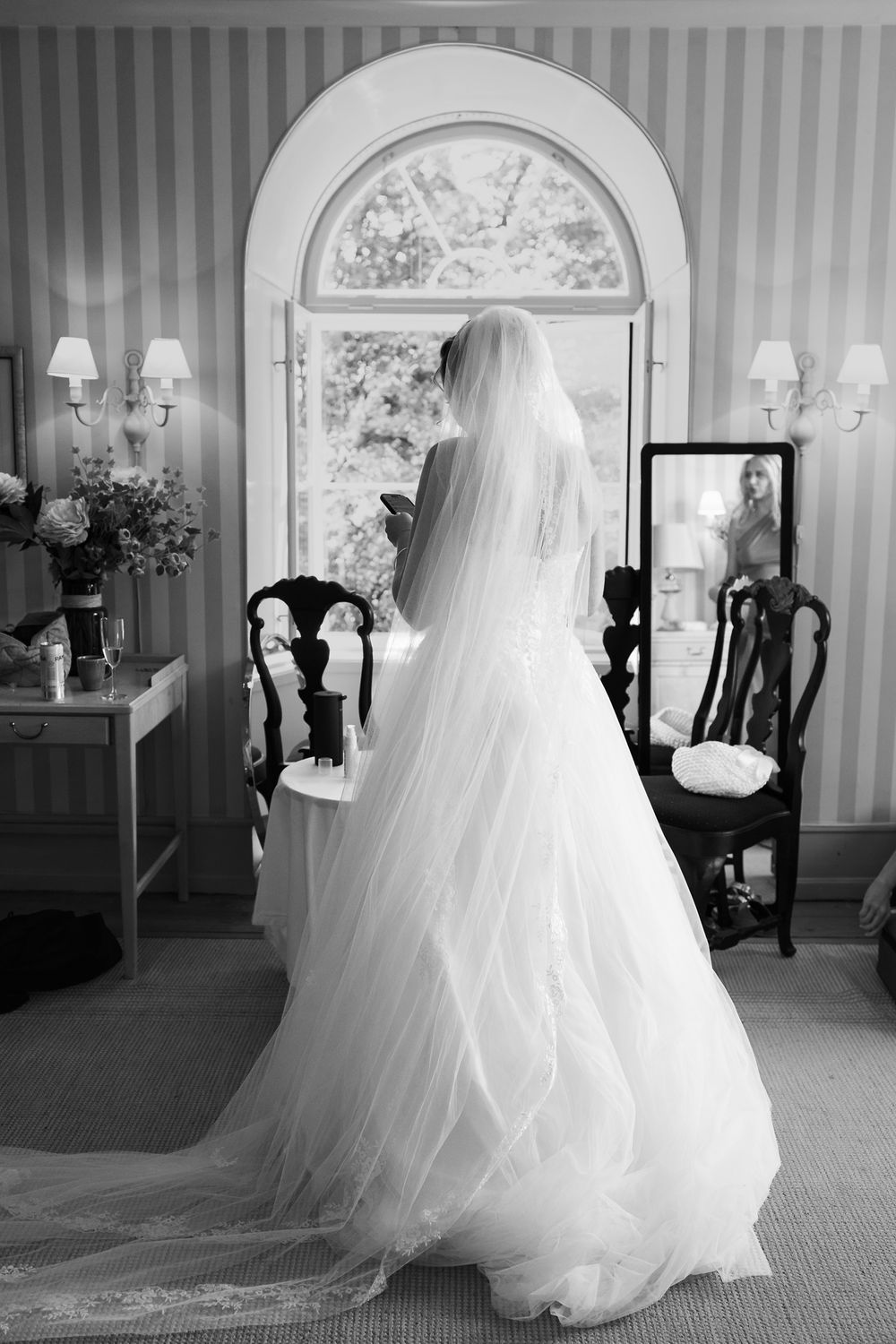 Bride in white gown and veil stands before arched window in elegant room, black and white wedding photo.