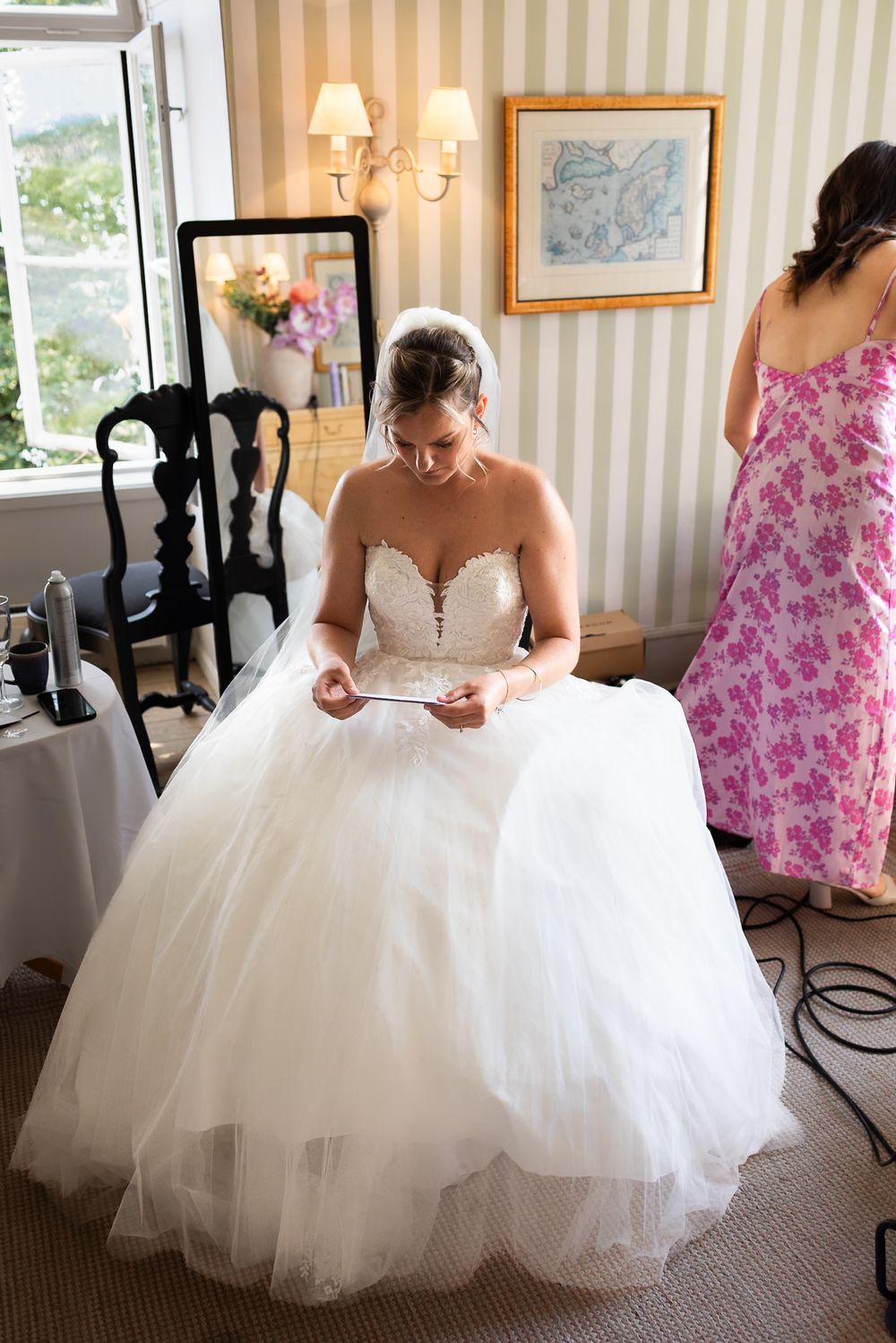 Bride in a white ballgown reads a note while a woman in a pink floral dress stands nearby in a bright room.