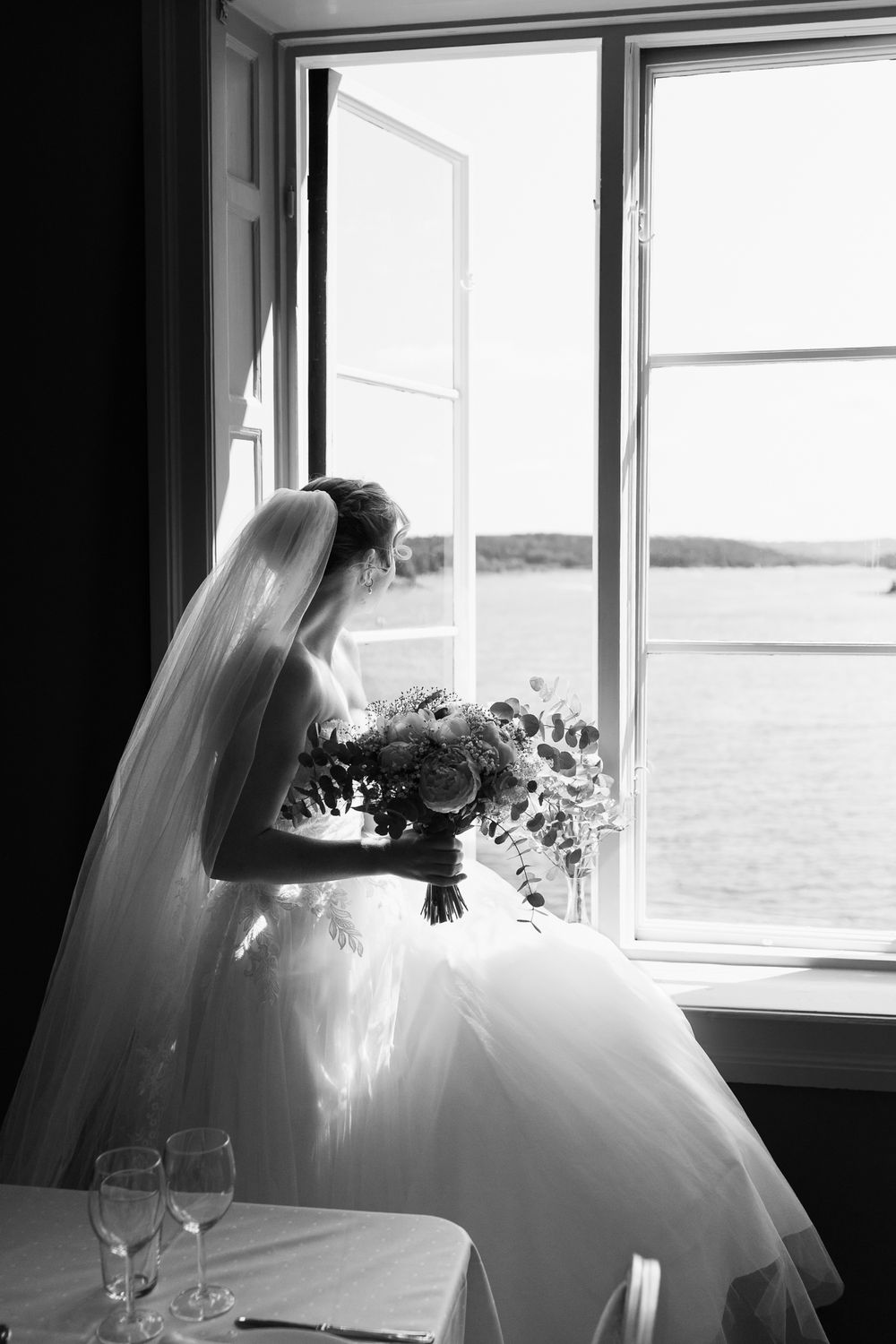 Black and white photo of a bride in a white gown holding a bouquet, gazing out an open window overlooking the sea.