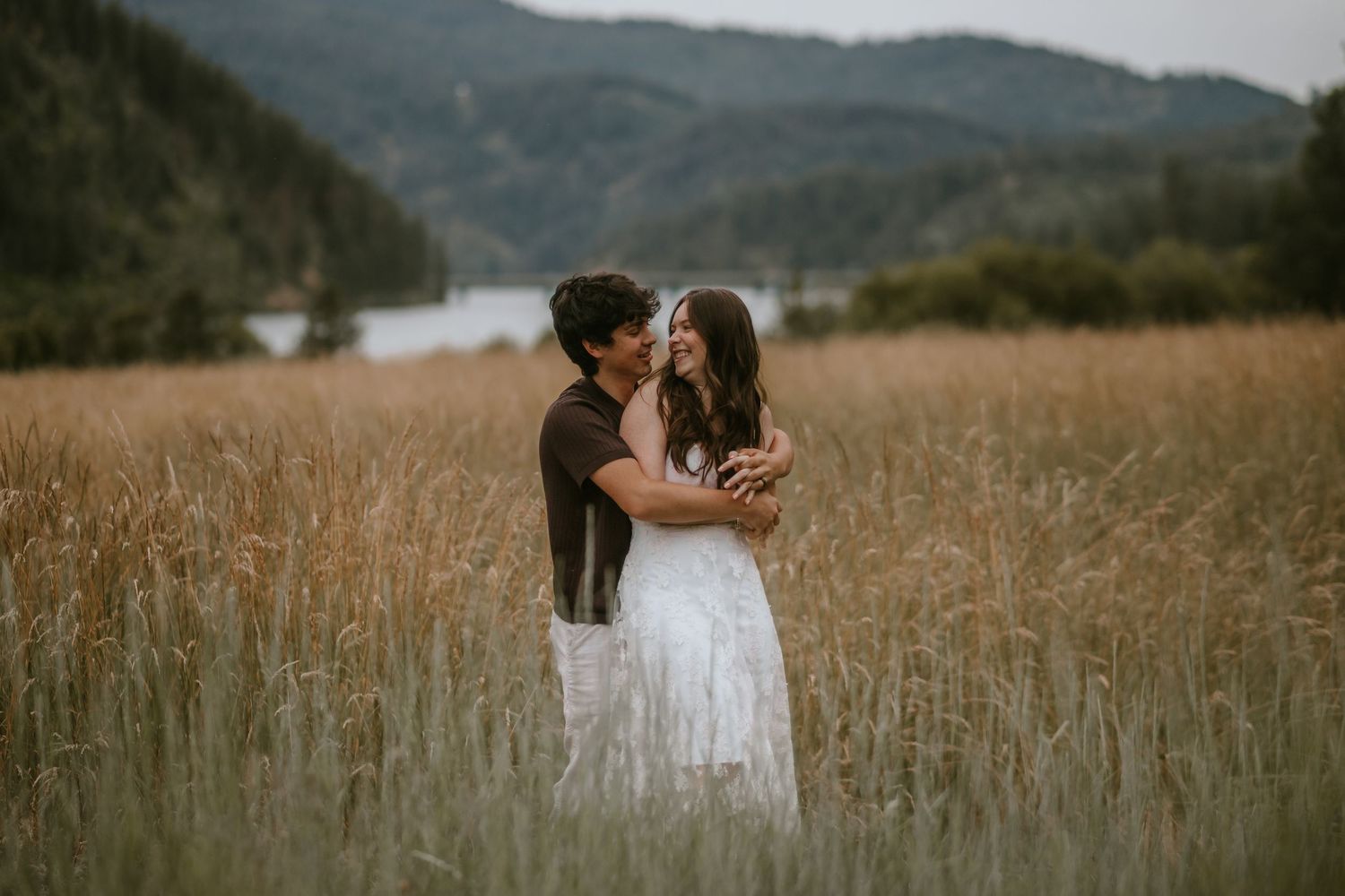 Couple embracing romantically in golden wheat field with mountain lake backdrop on overcast day.