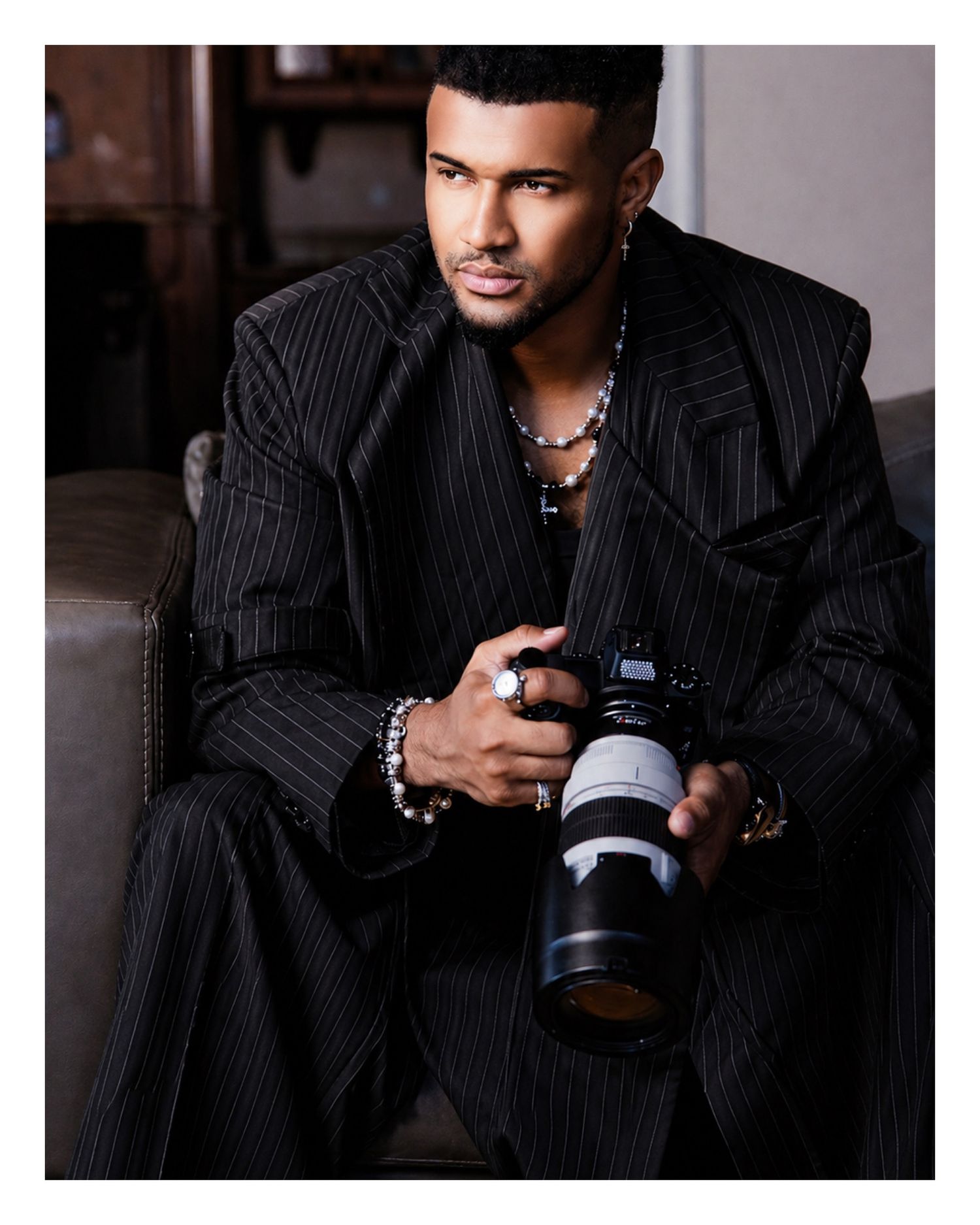 A stylish man in a black pinstripe suit holds a professional camera with a telephoto lens while seated on a sofa.