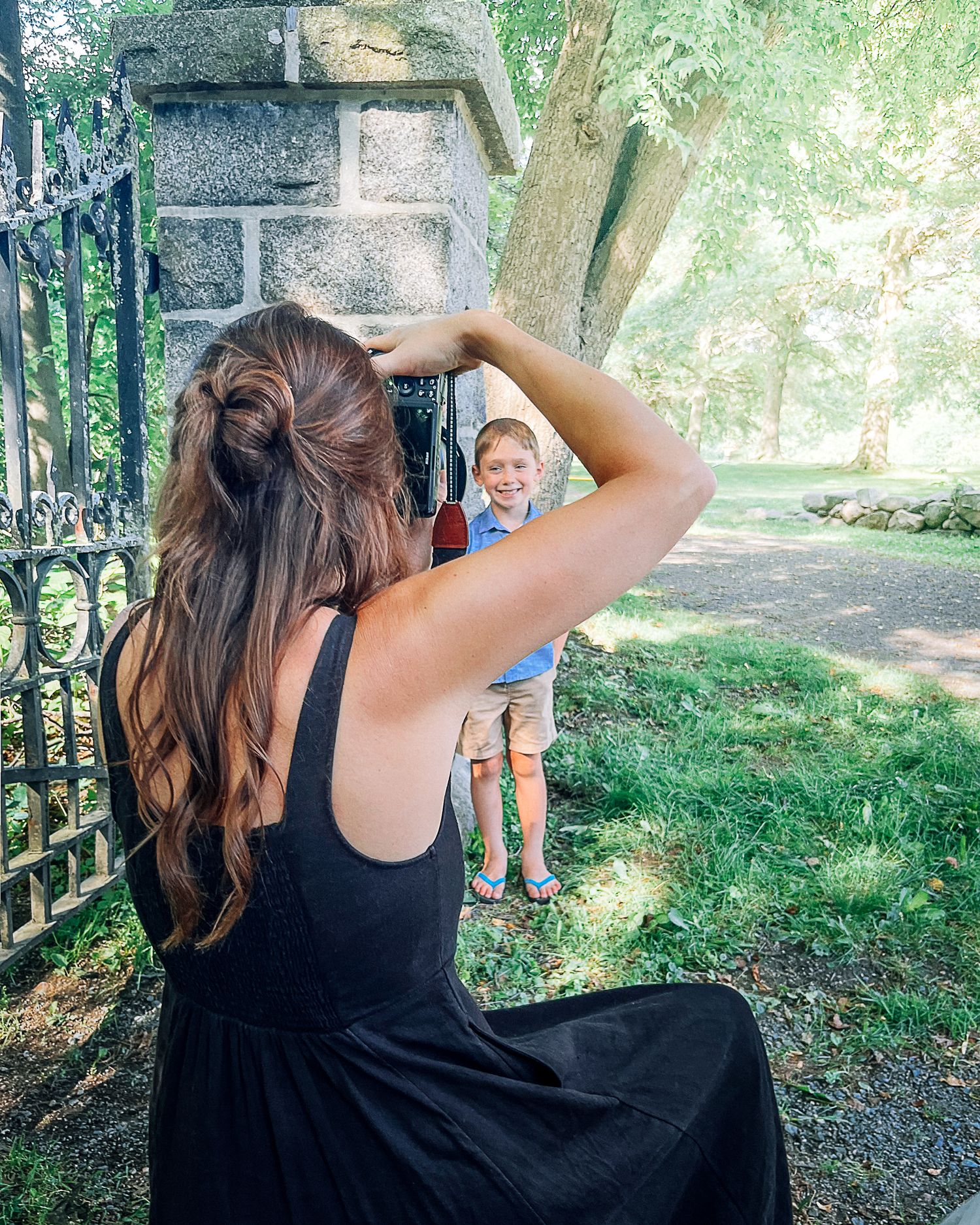 Woman in black dress photographing a smiling boy near ornate iron gates in a lush green park.