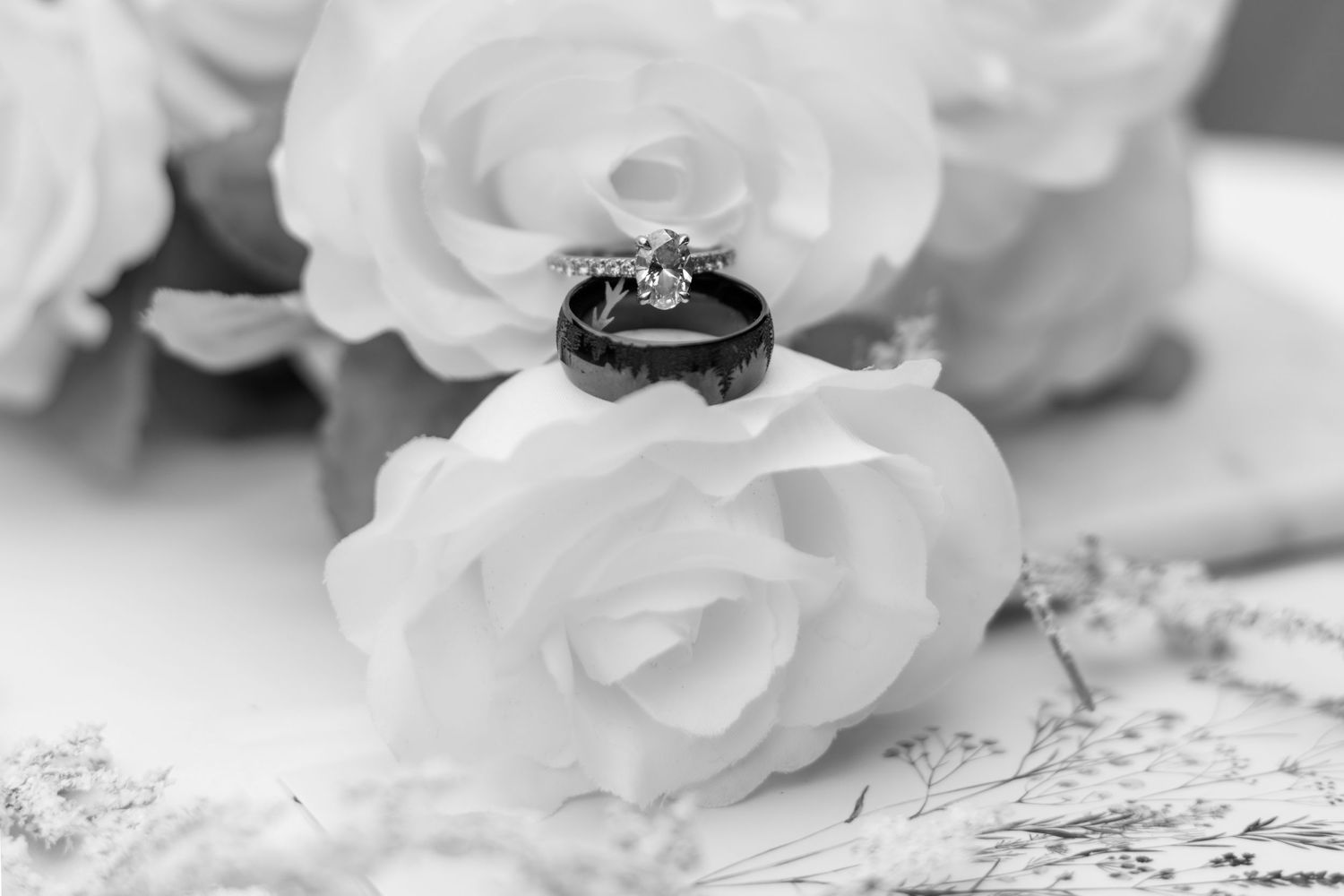 Black and white photo of wedding rings resting on white roses with elegant floral background.