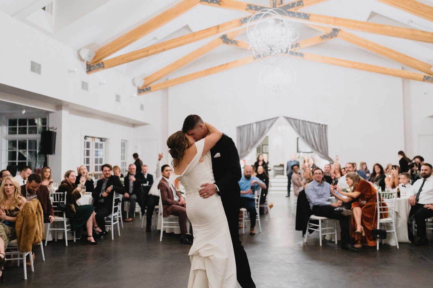 Bride and groom share first dance at elegant white wedding reception venue with exposed beam ceilings and seated guests.