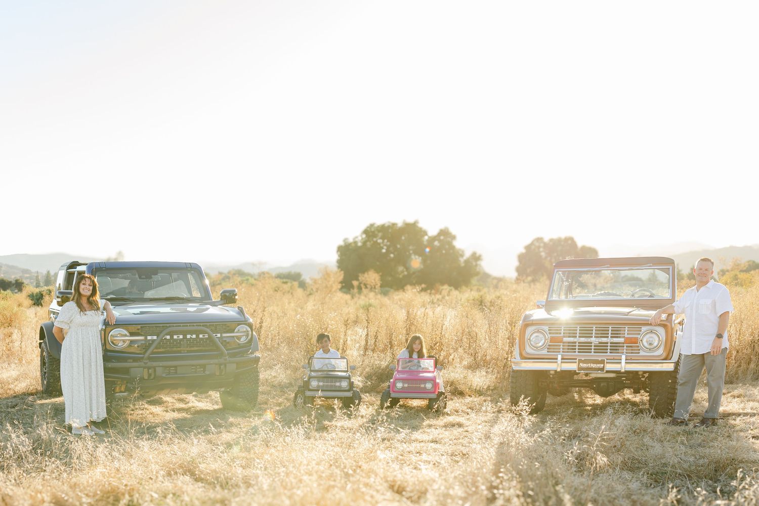 Family posing in golden field between a new Ford Bronco and vintage Bronco during sunny outdoor photo session.