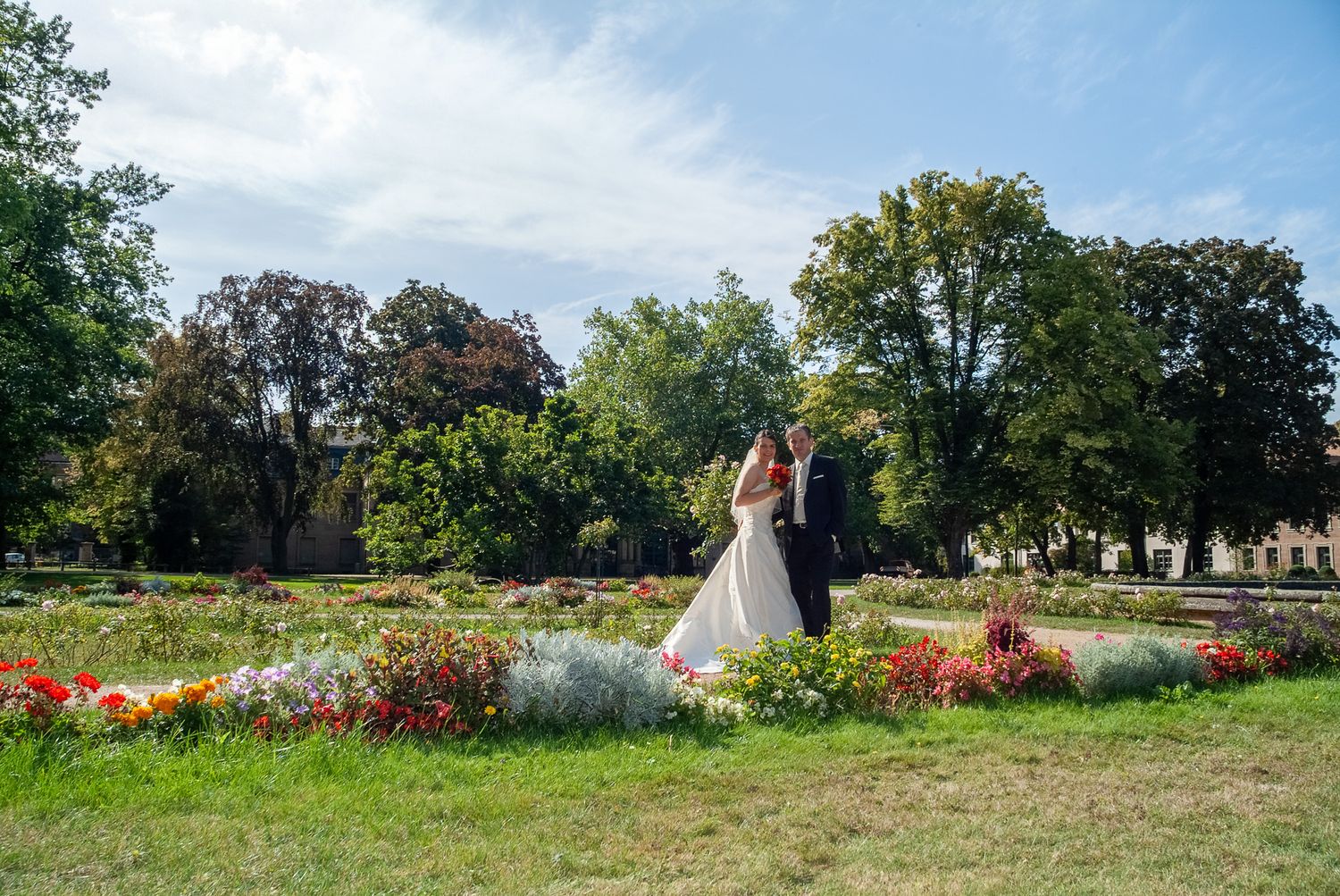 Brautpaar in der Ferne hinter bunten Blumenbeeten im Park. Weitläufige Hochzeitsfotografie durch die Lichtkapsel in Erlangen.