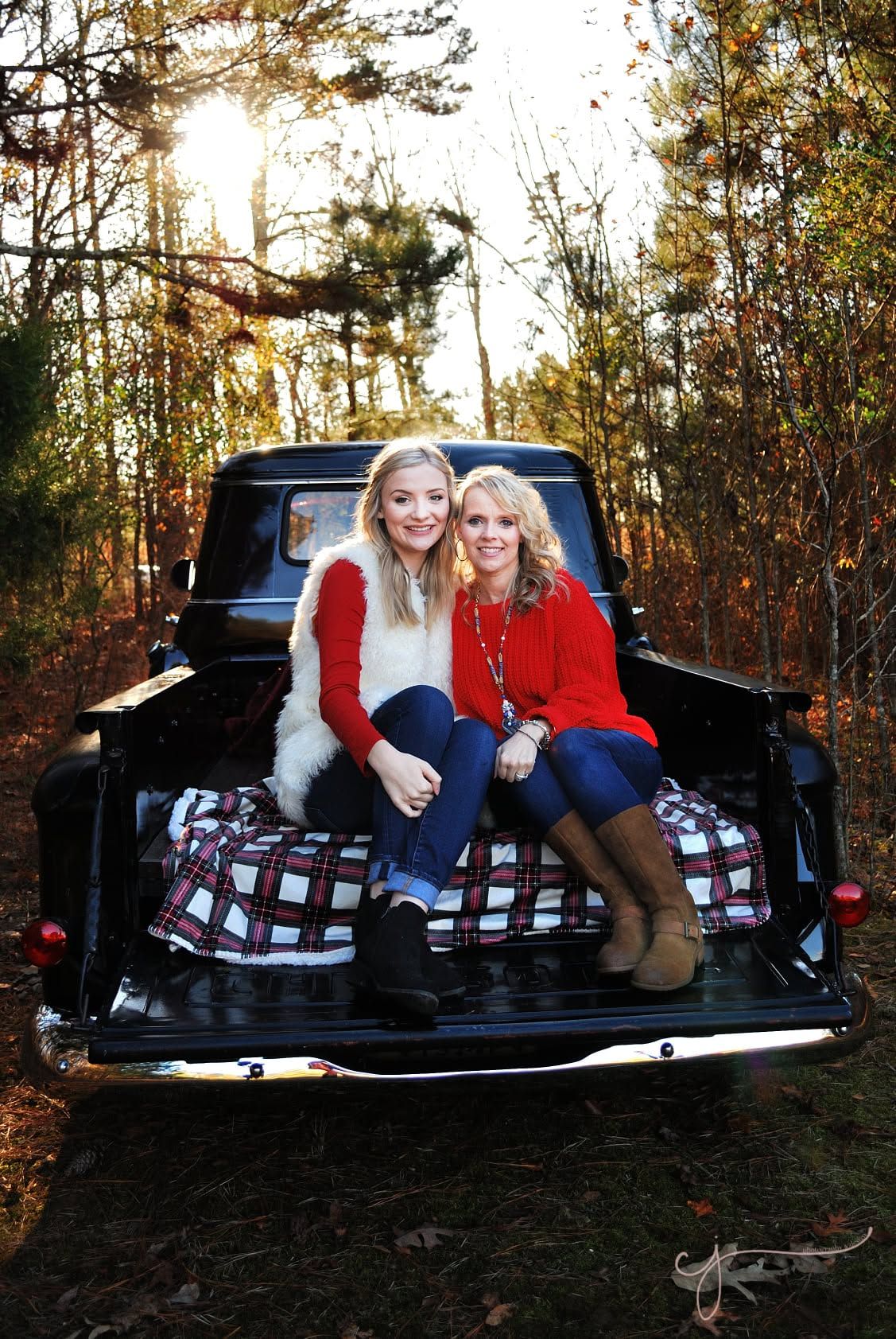 Two women in red and white outfits sit smiling on a plaid blanket in a vintage black truck bed surrounded by autumn trees.