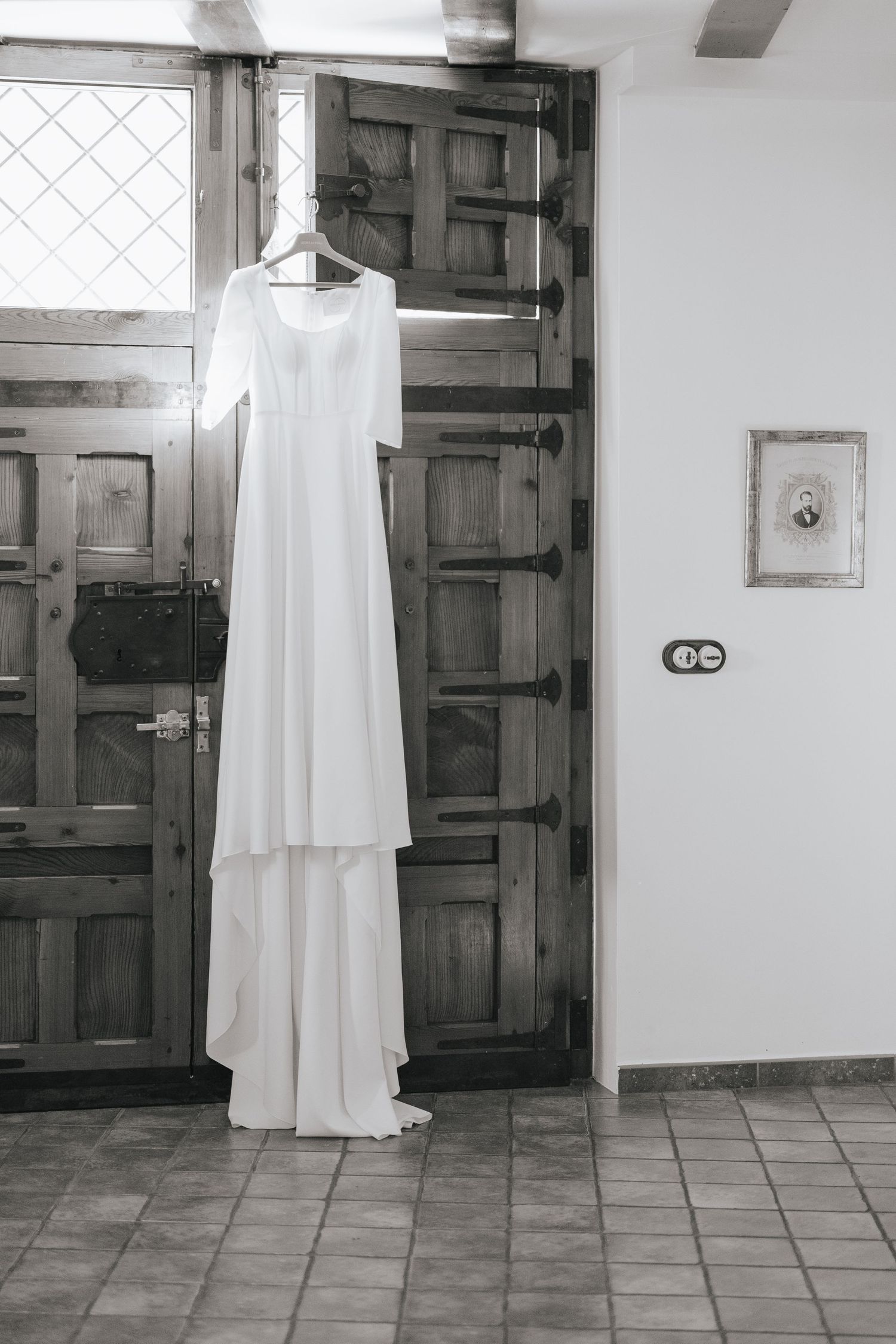 Black and white photo of an elegant white wedding dress hanging on a wooden door in a tiled room.