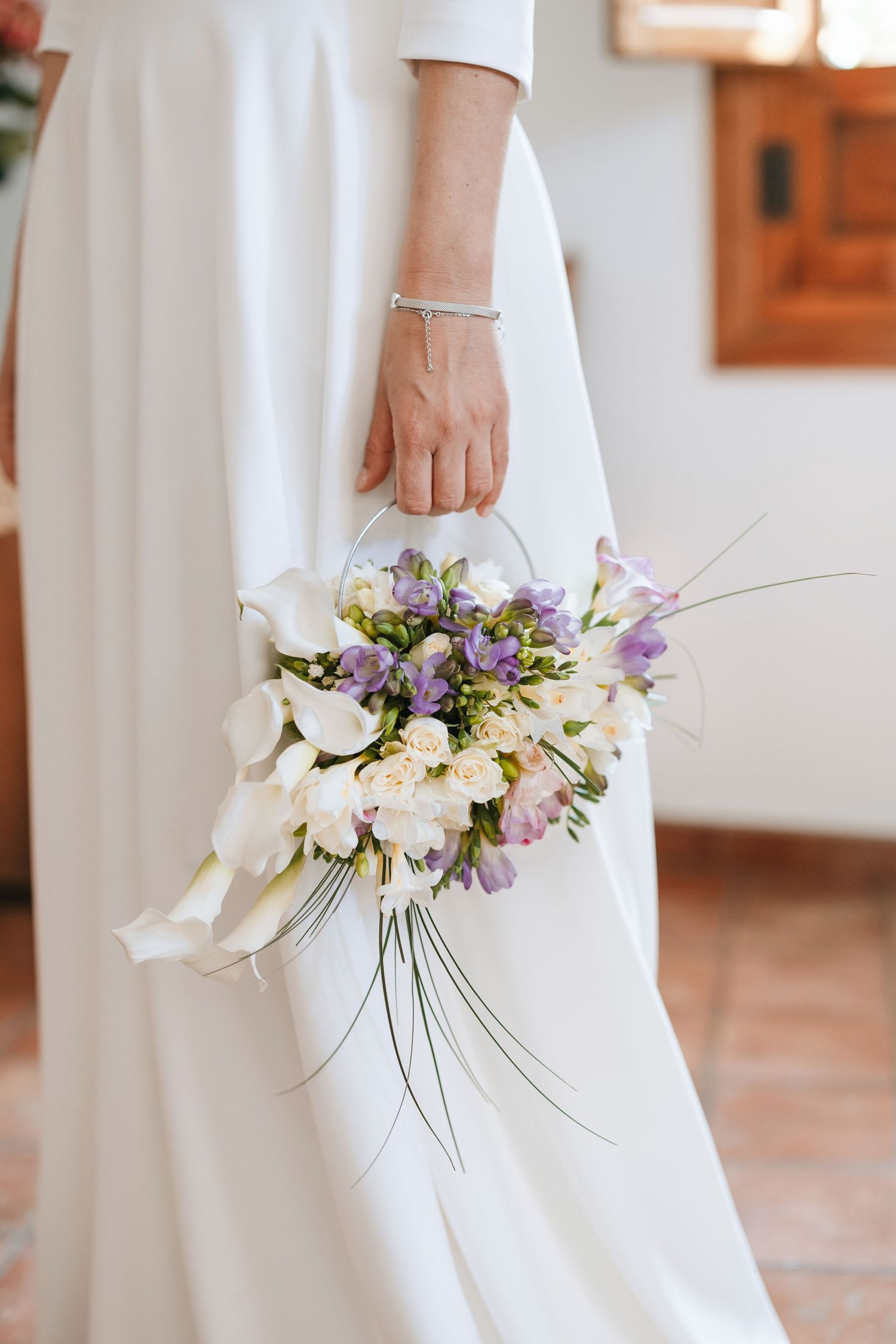 Bride in white gown holding elegant bouquet of white calla lilies and purple freesias with trailing grass accents.