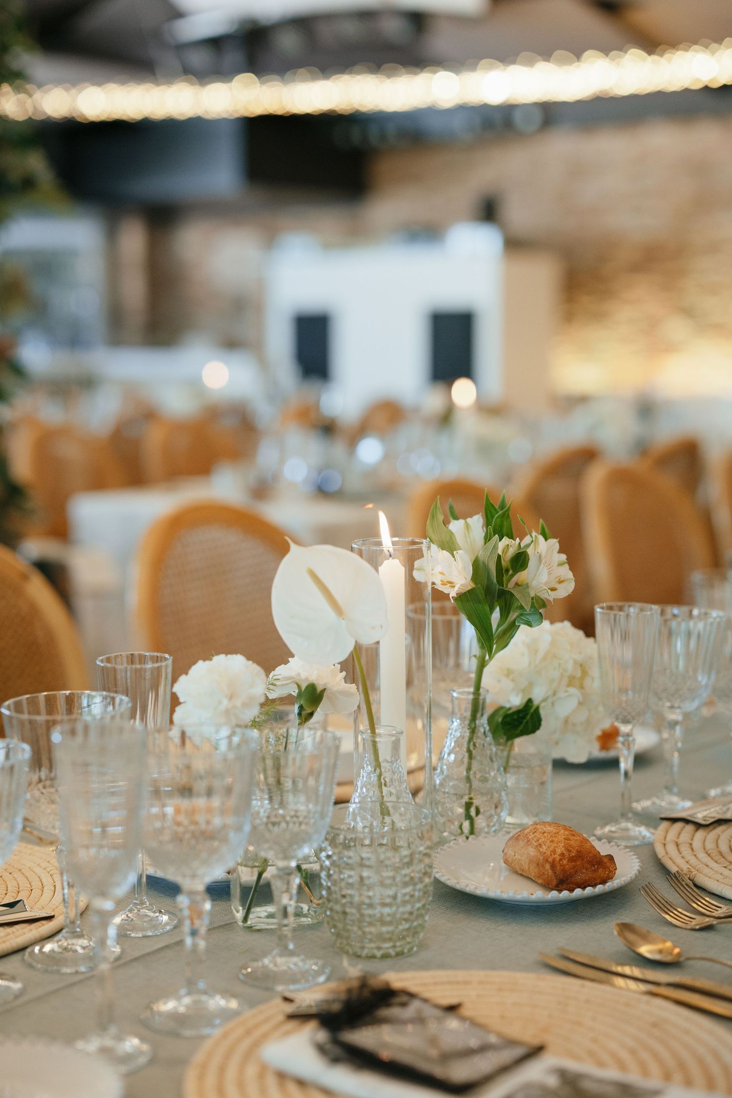 Elegant wedding reception table with white floral centerpiece, crystal glasses, candles, and wicker chairs in soft lighting.