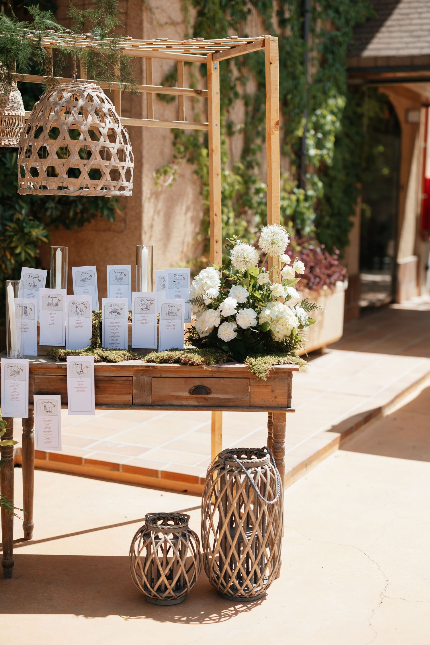 Rustic wooden table with white floral arrangements, wicker lanterns, and place cards at an outdoor wedding venue.