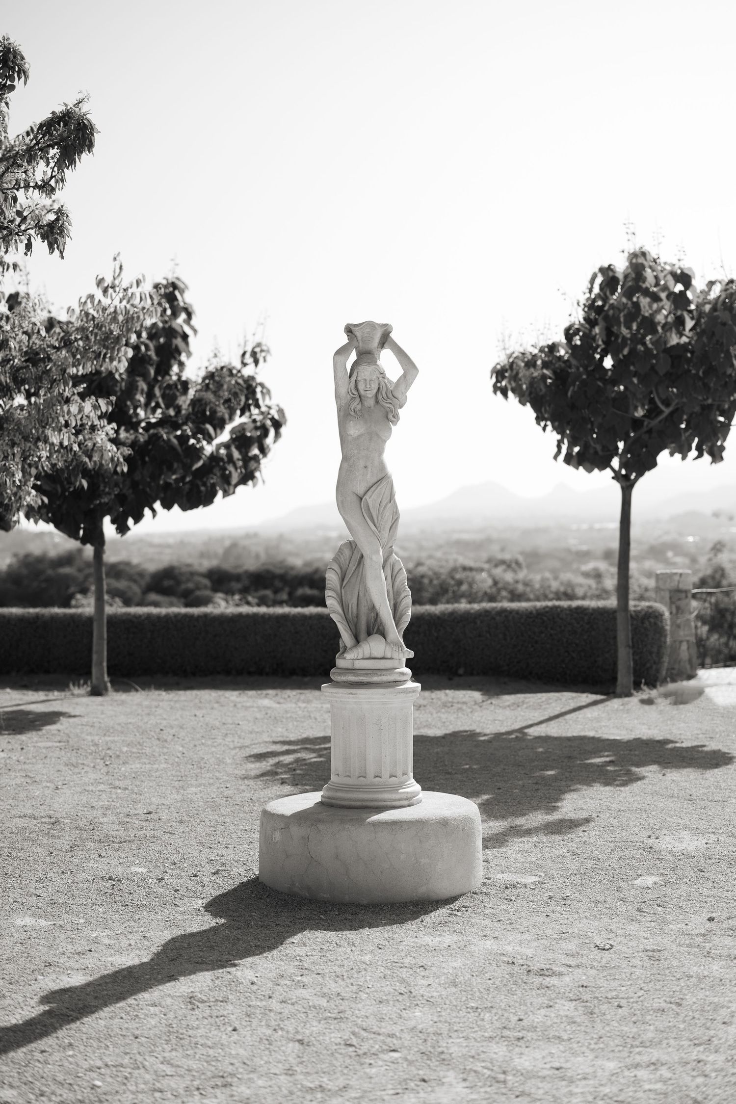 Black and white photo of a classical female statue on a pedestal in a formal garden with trimmed trees.