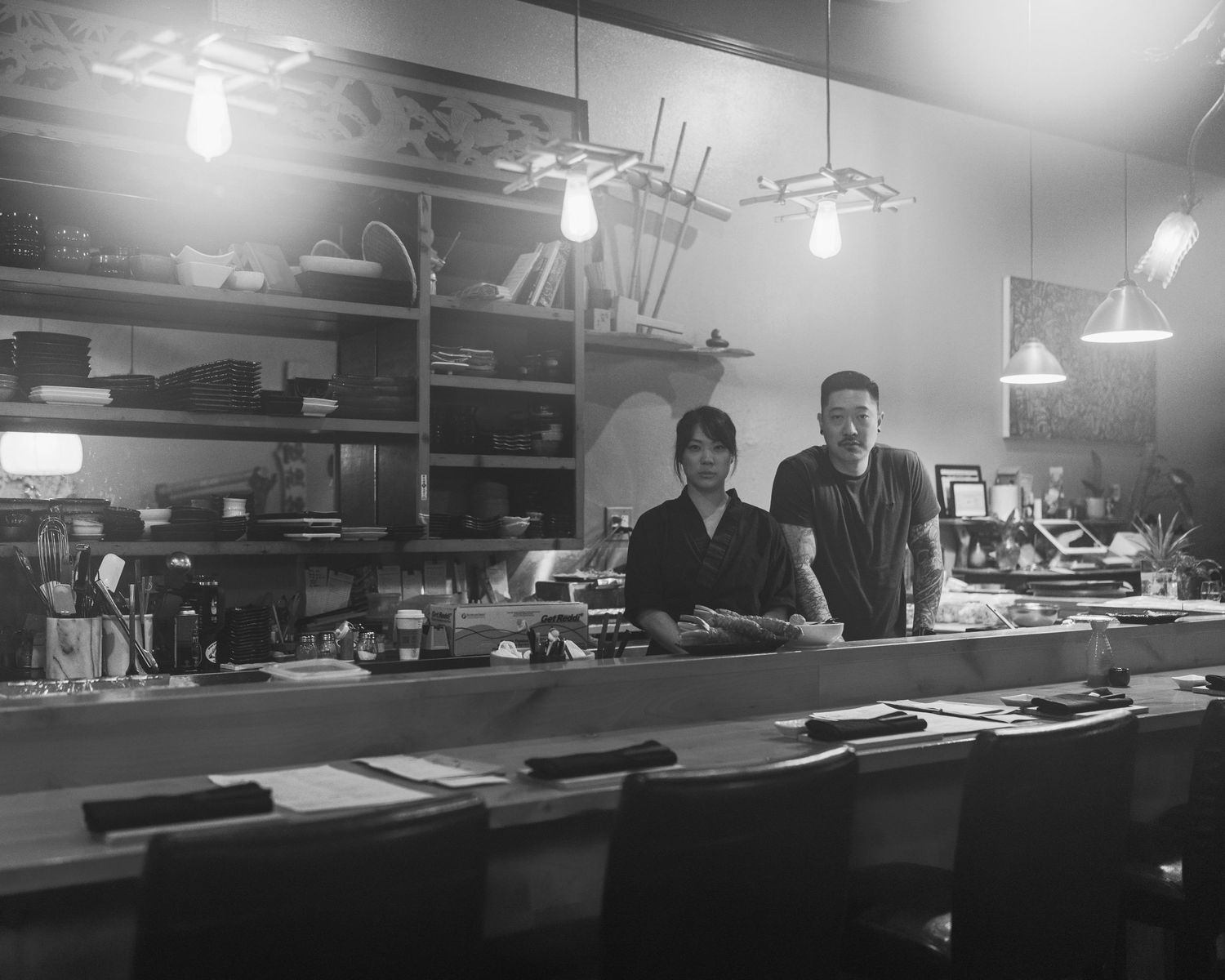 Two staff members stand behind a sushi bar counter in a warmly lit Japanese restaurant, black and white photo.