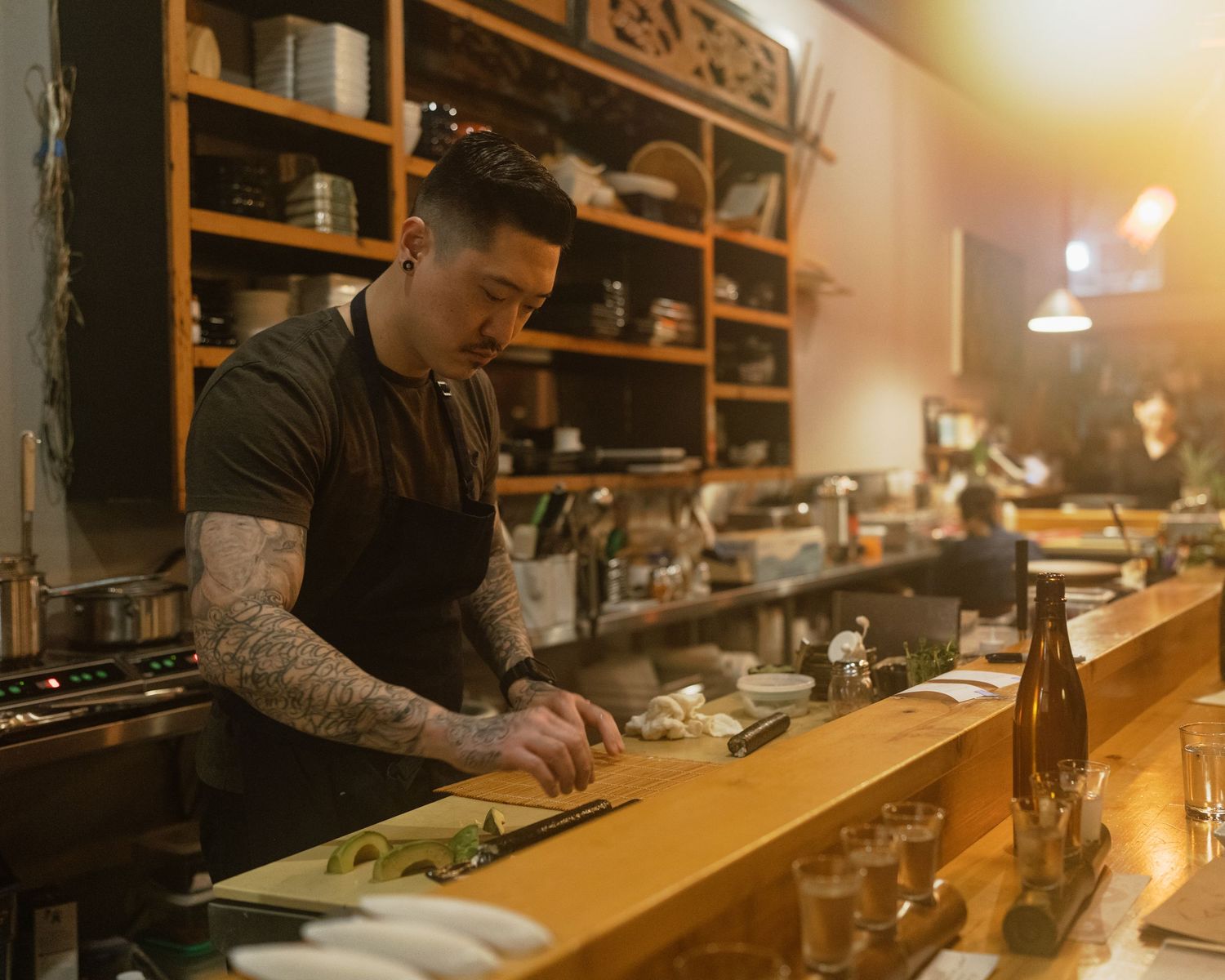 Tattooed chef in black apron prepares food at wooden counter in a warmly lit Japanese restaurant kitchen.
