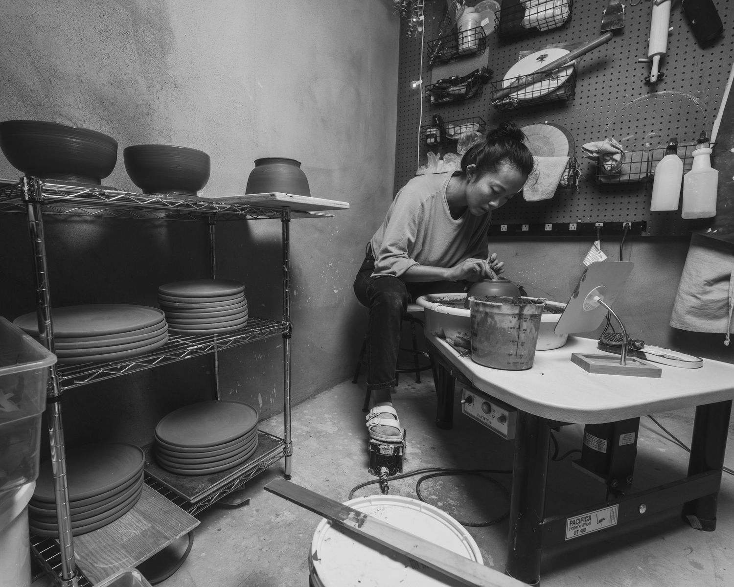 Black and white photo of a person working at a pottery wheel in a ceramic studio with shelves of plates nearby.