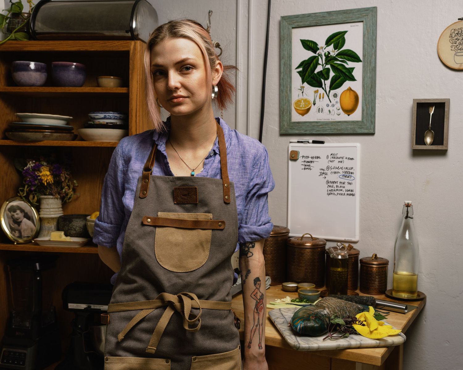 Young woman in gray canvas apron stands confidently in a rustic kitchen with botanical art and jars.